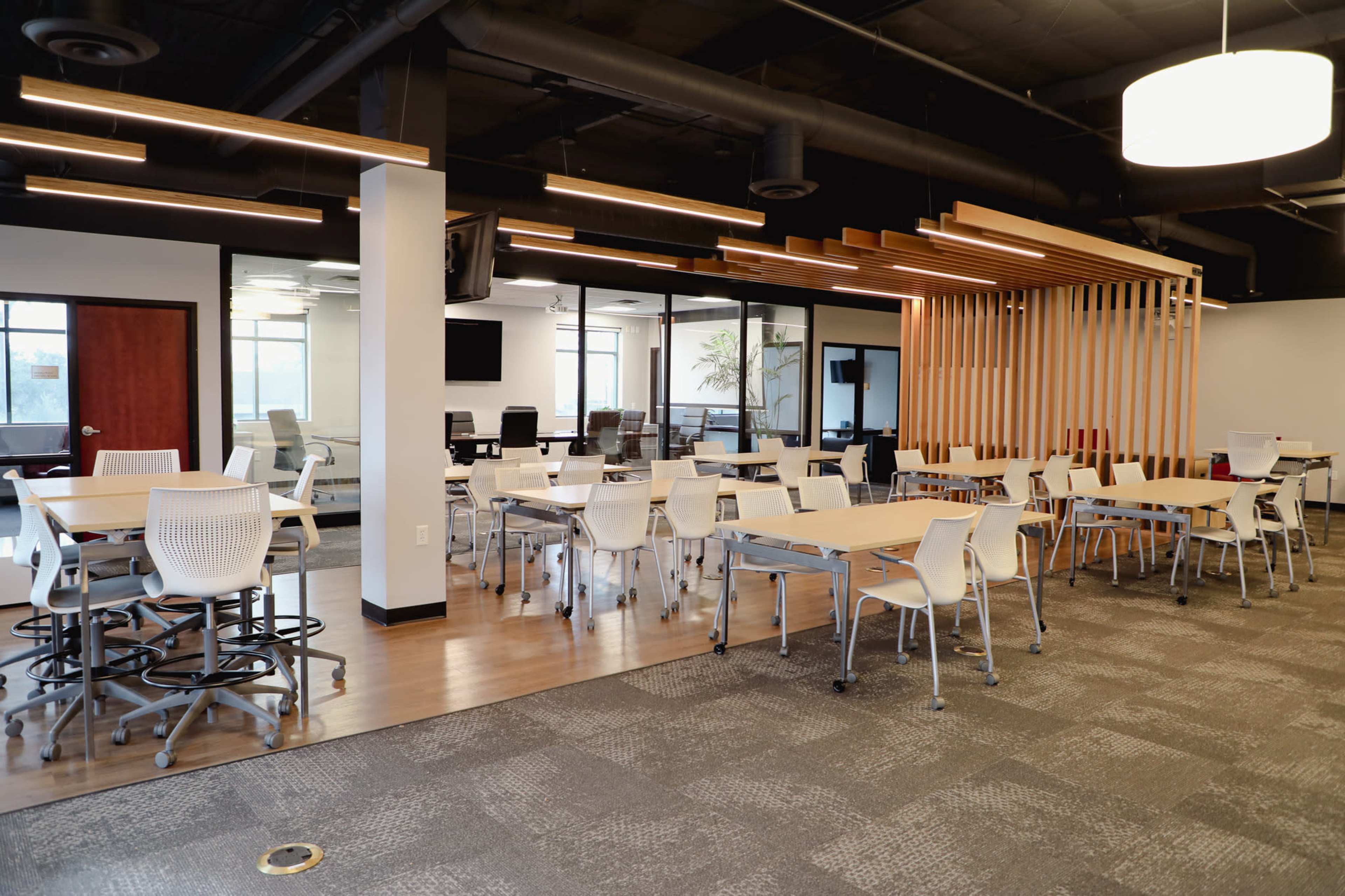 A modern office space with several tables and chairs arranged for collaborative work, along with glass-walled meeting rooms in the background.