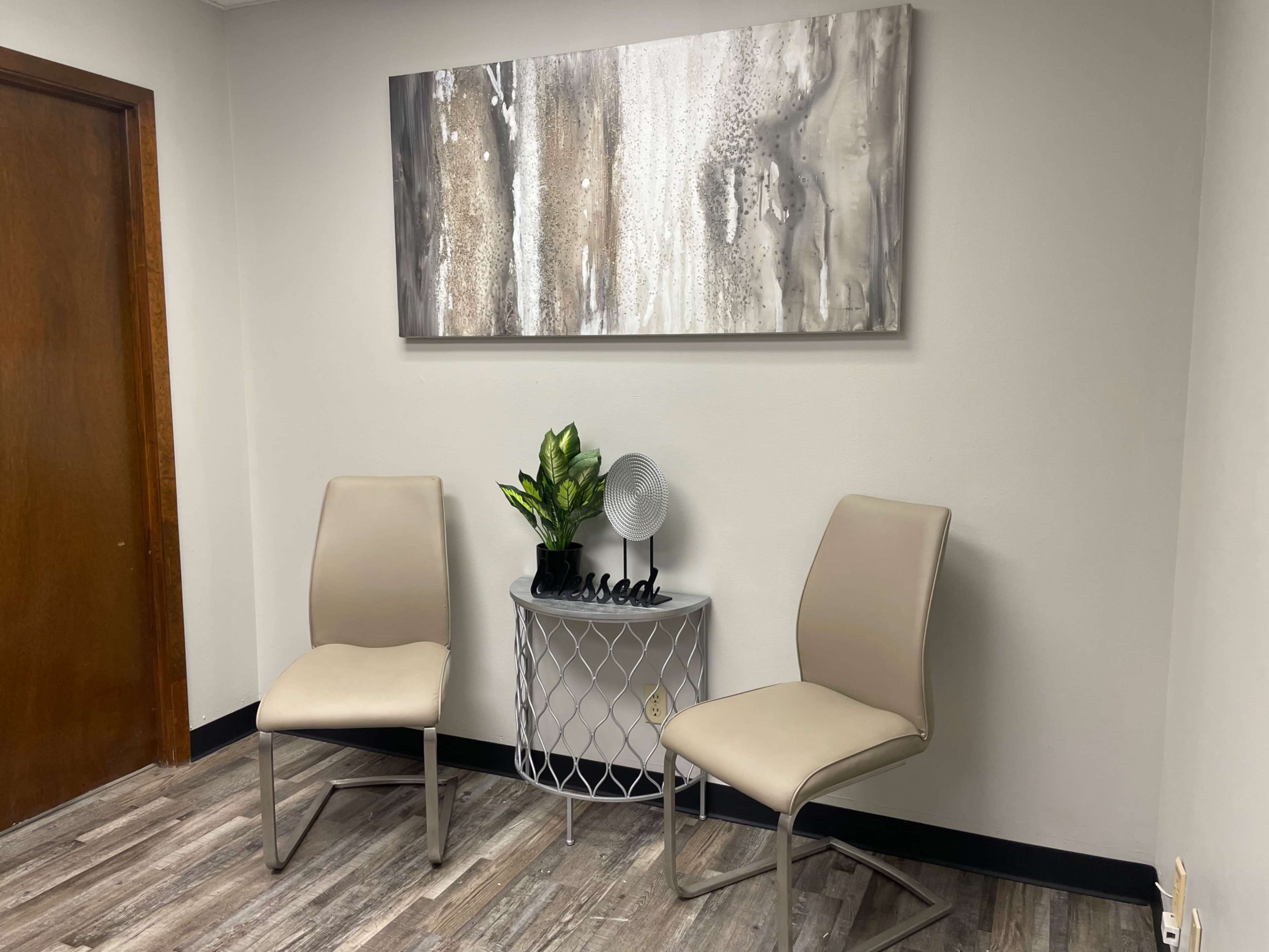 The image shows a minimalist waiting area with two beige chairs, a small round table, a decorative plant, and a fan against a neutral wall, featuring an abstract painting above.