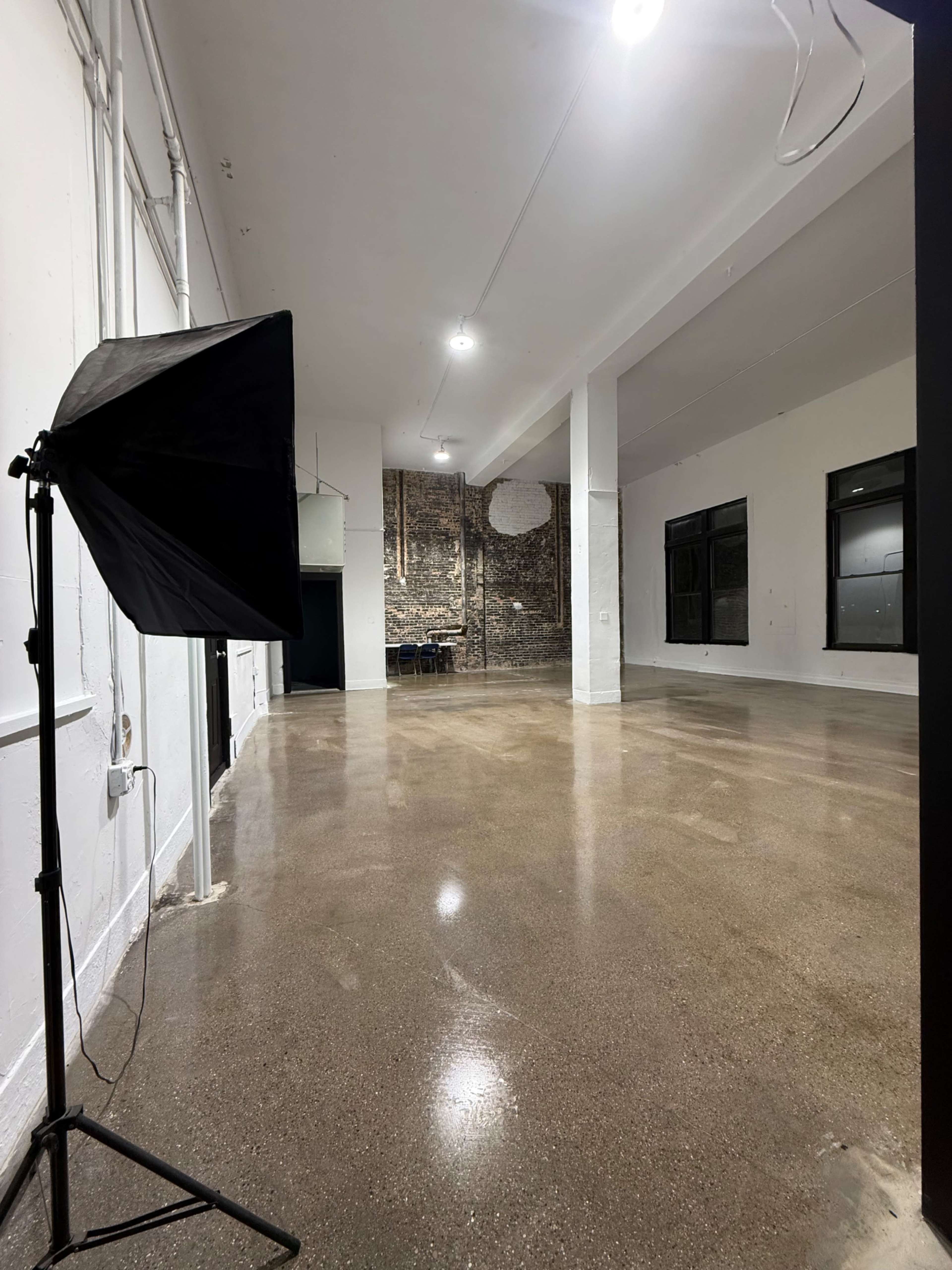 The image shows a spacious, well-lit room with polished concrete floors, a brick wall, and a photography light stand positioned in one corner.