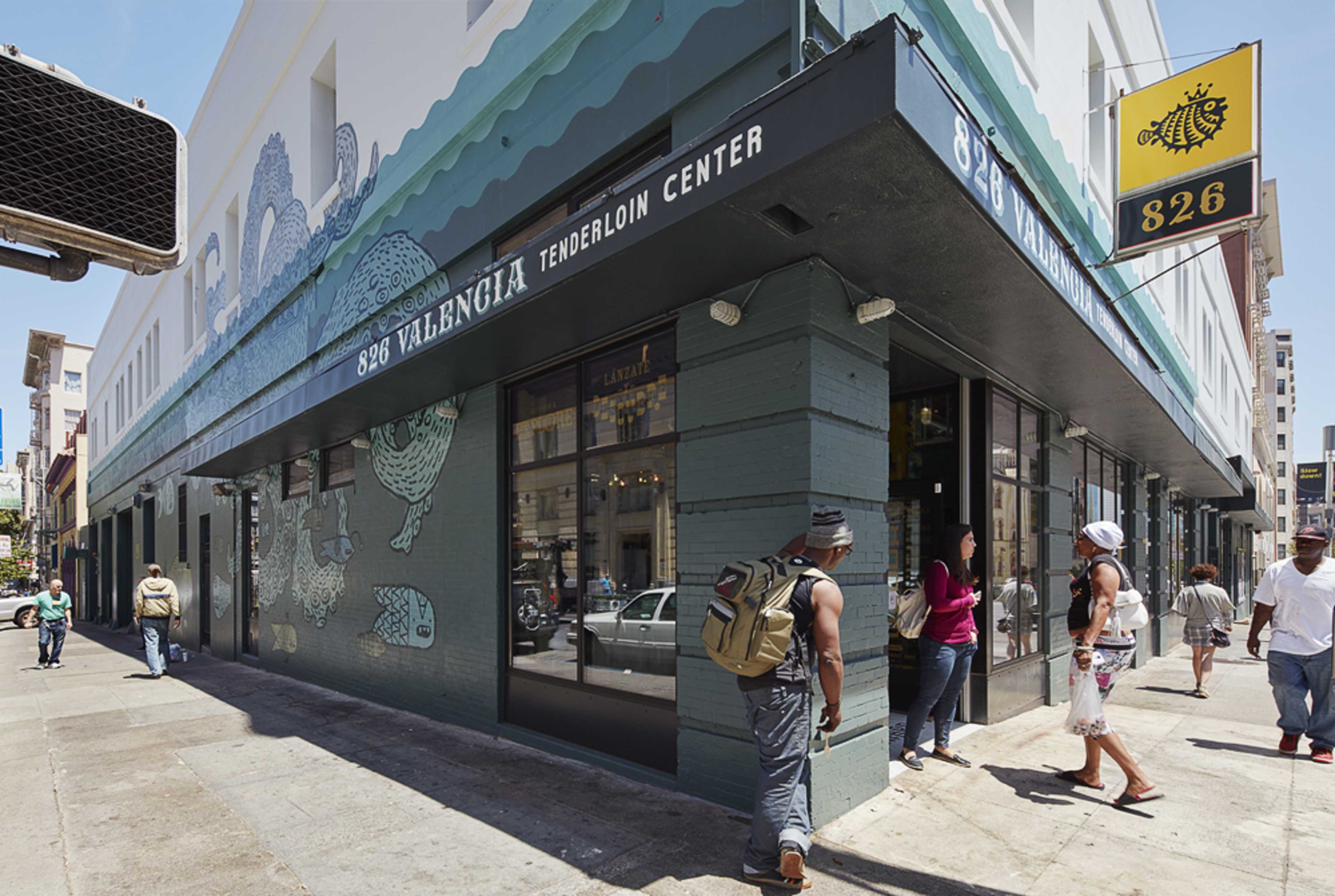 A colorful building exterior with a sign identifying the Tenderloin Center at 580 Valencia Street, featuring several pedestrians and storefronts in a city setting.