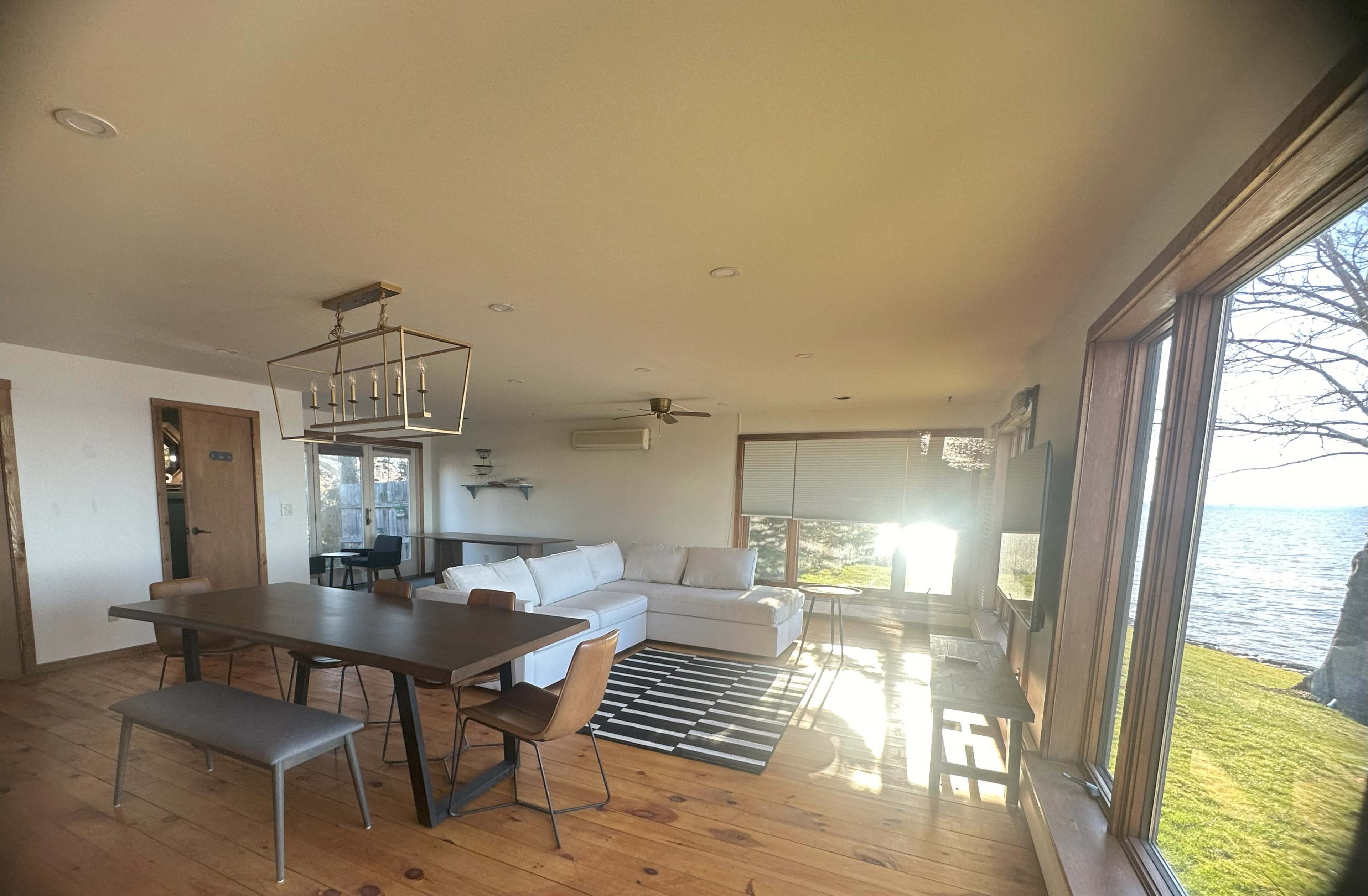 A bright living space with a dining area, a sofa, and large windows overlooking a body of water.