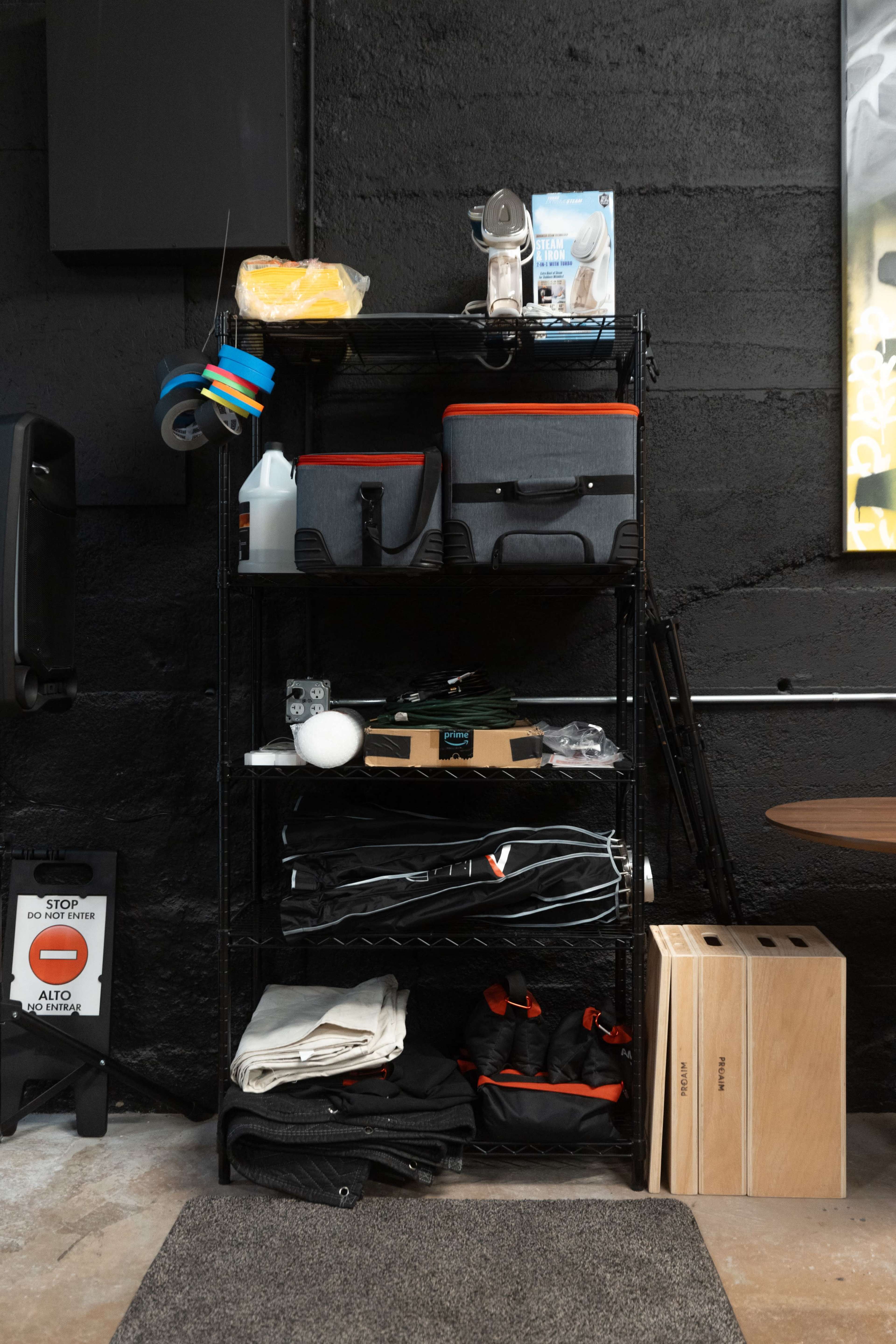 A black metal shelving unit filled with storage bins, cleaning supplies, and rolled fabric arranged neatly against a textured wall.