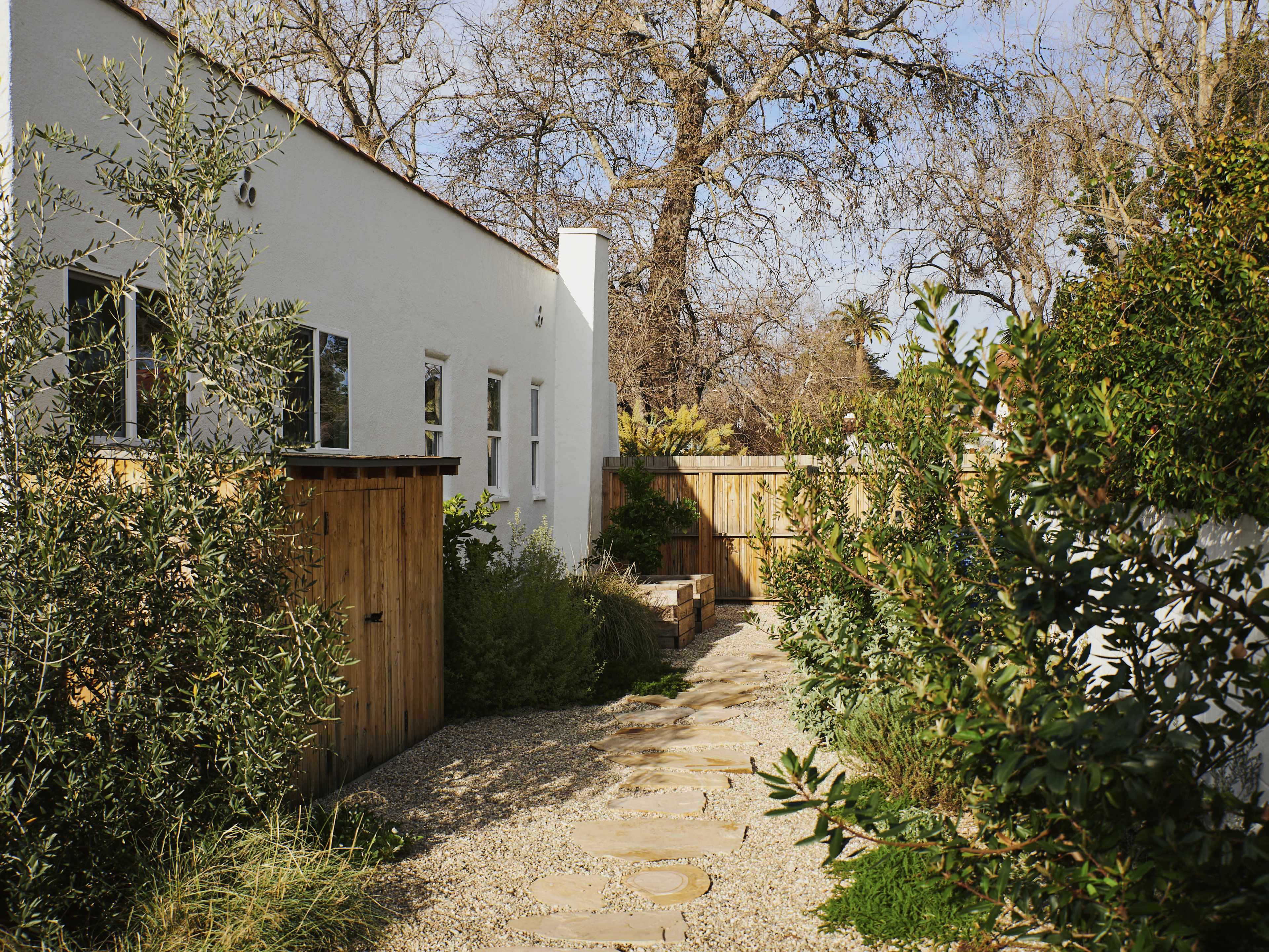 A stone path lined with greenery leads to a wooden gate beside a white building set in a tranquil outdoor space.