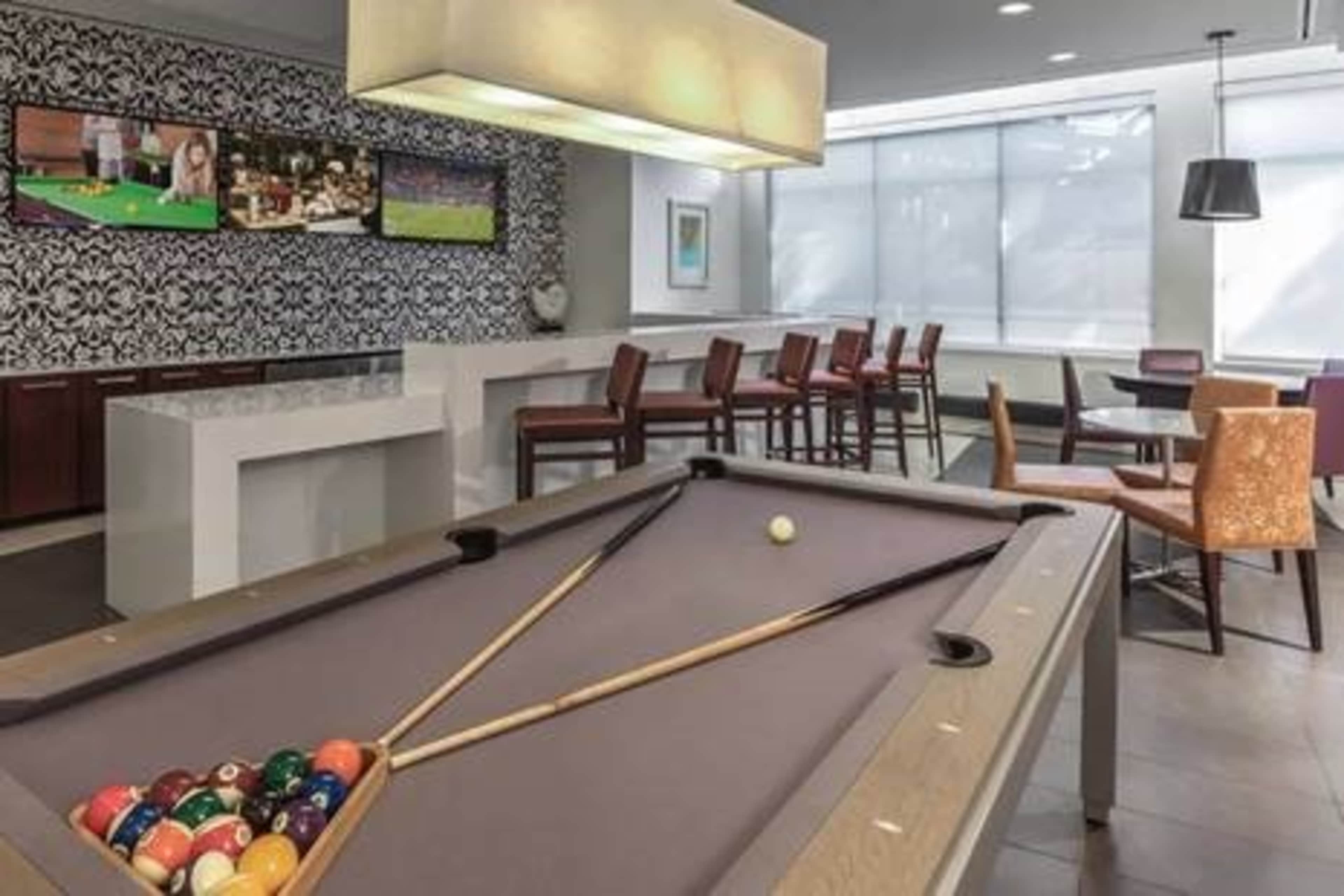 A modern recreational room featuring a pool table with a triangle rack of balls, surrounded by tables and chairs under bright lighting.