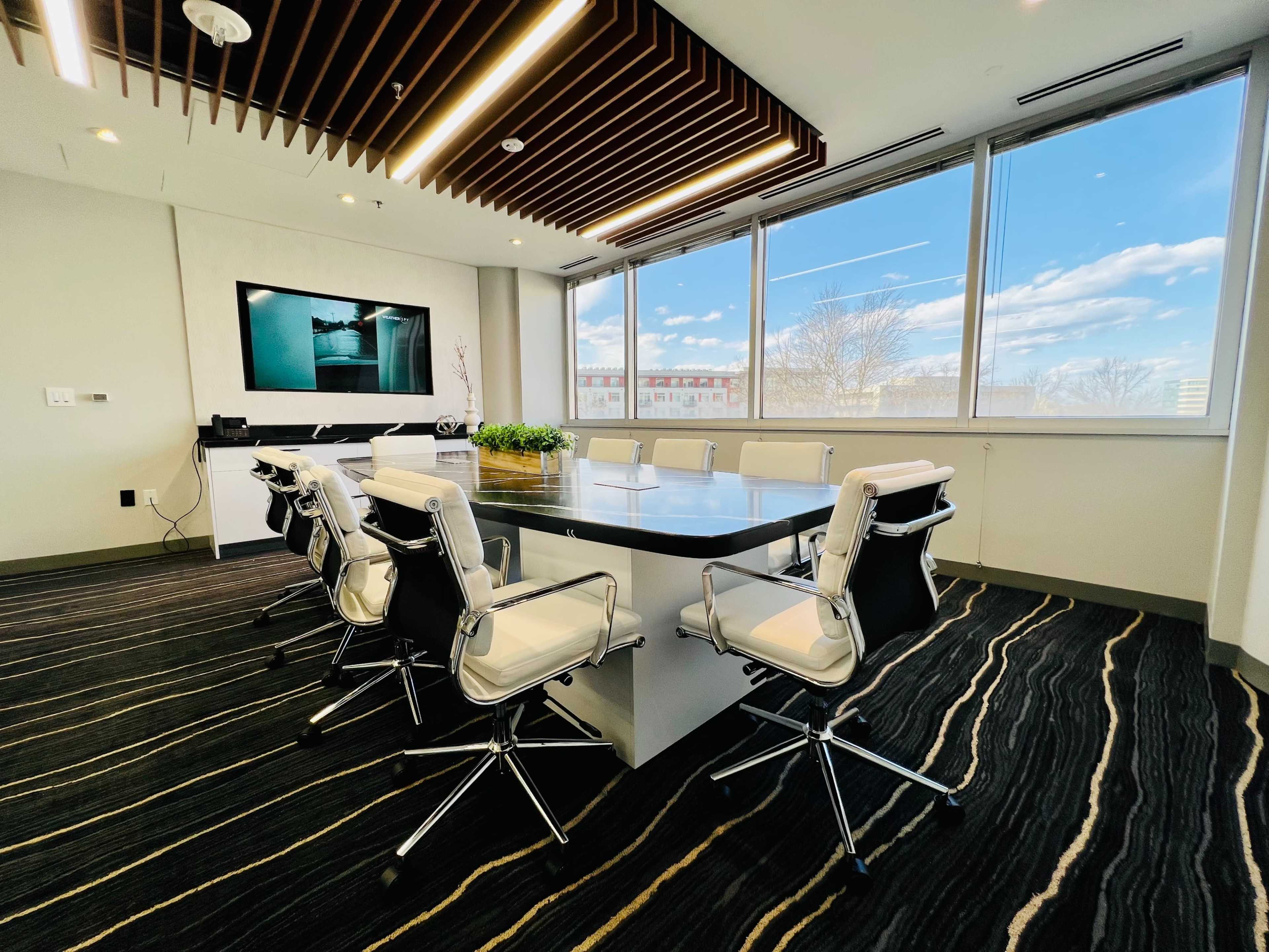 A modern conference room features a large table surrounded by white leather chairs, with a television mounted on the wall and large windows displaying a cloudy sky.