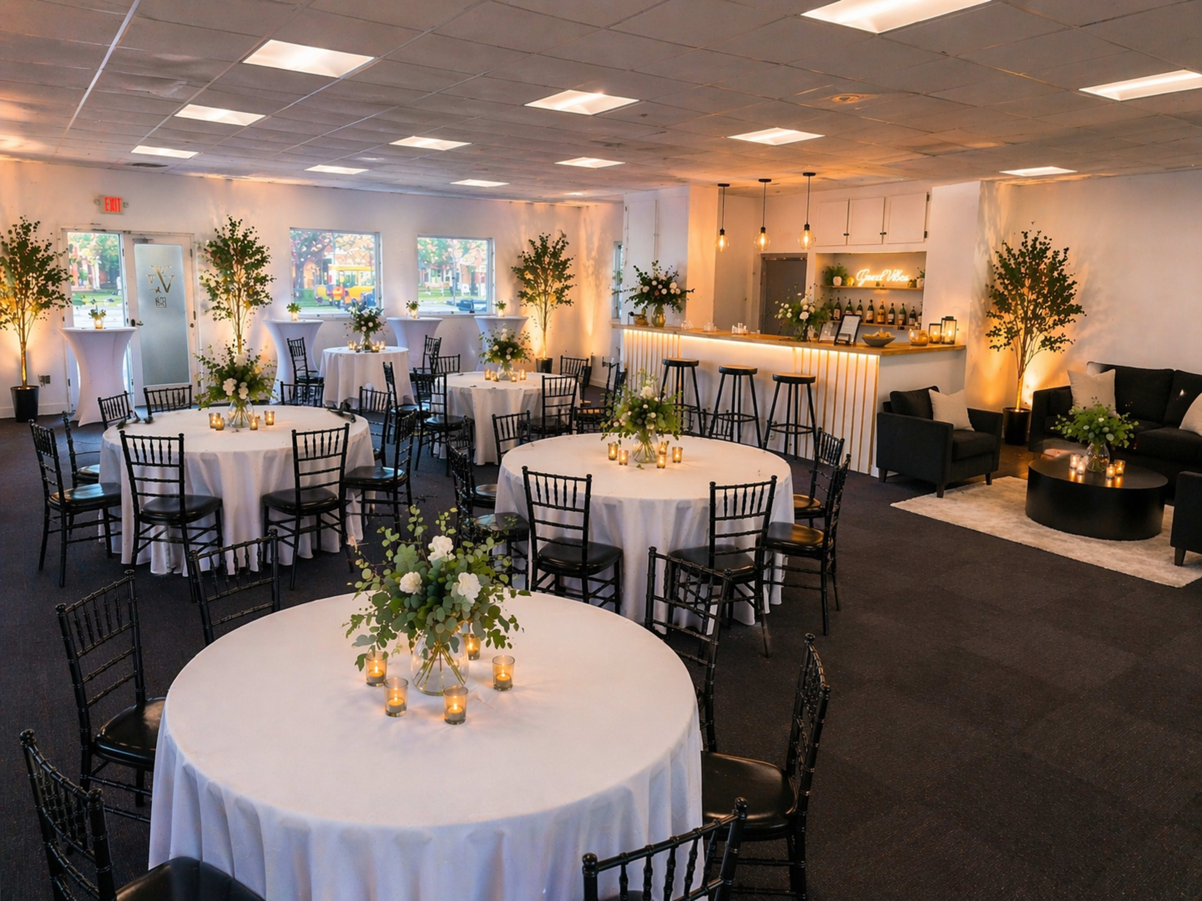The image shows a banquet space with round tables set for an event, featuring floral centerpieces, illuminated by soft lighting from the bar area in the background.