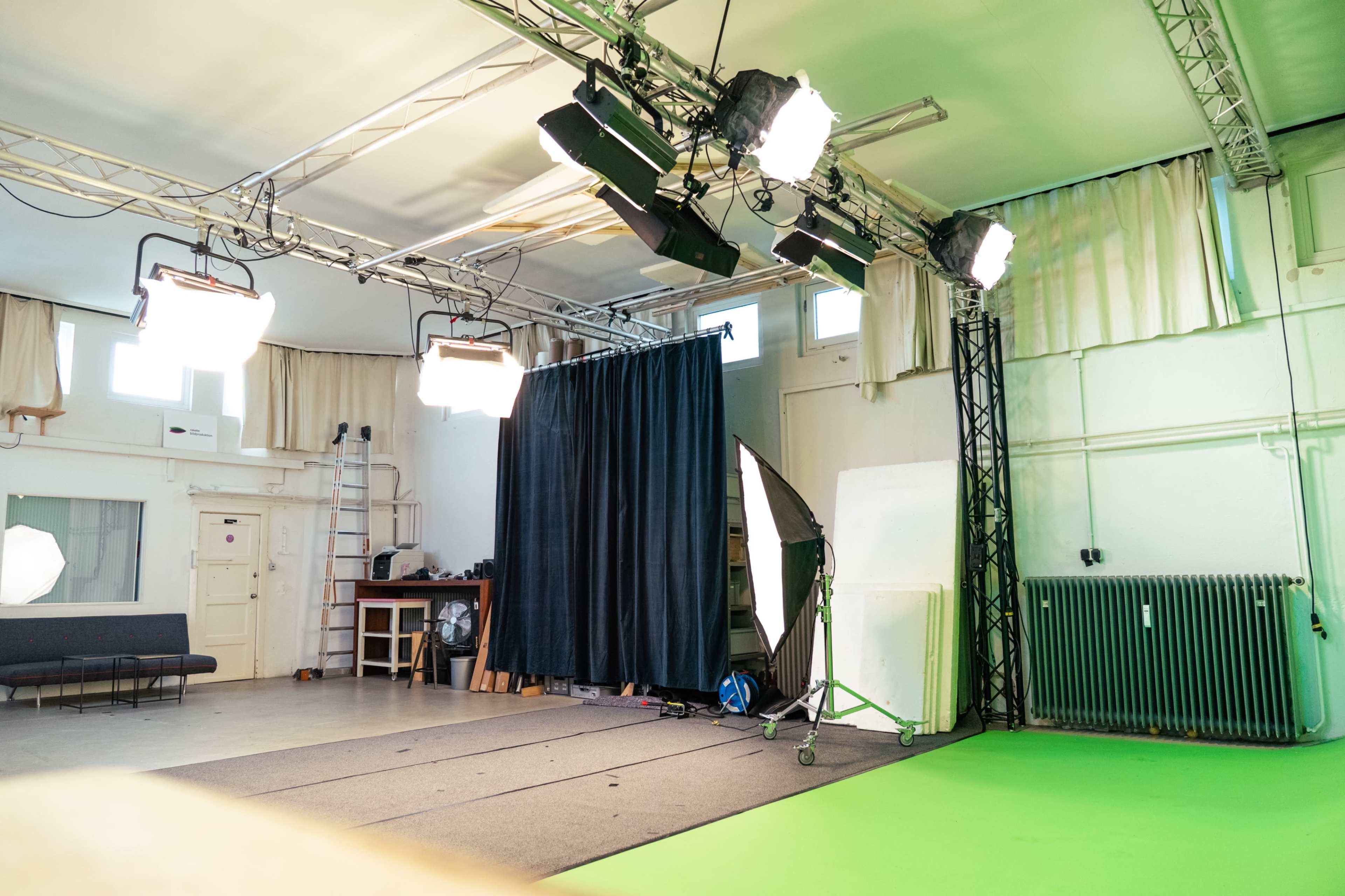 The image shows a photography studio with green flooring, lighting rigs, a black curtain, and various equipment set up for a shoot.