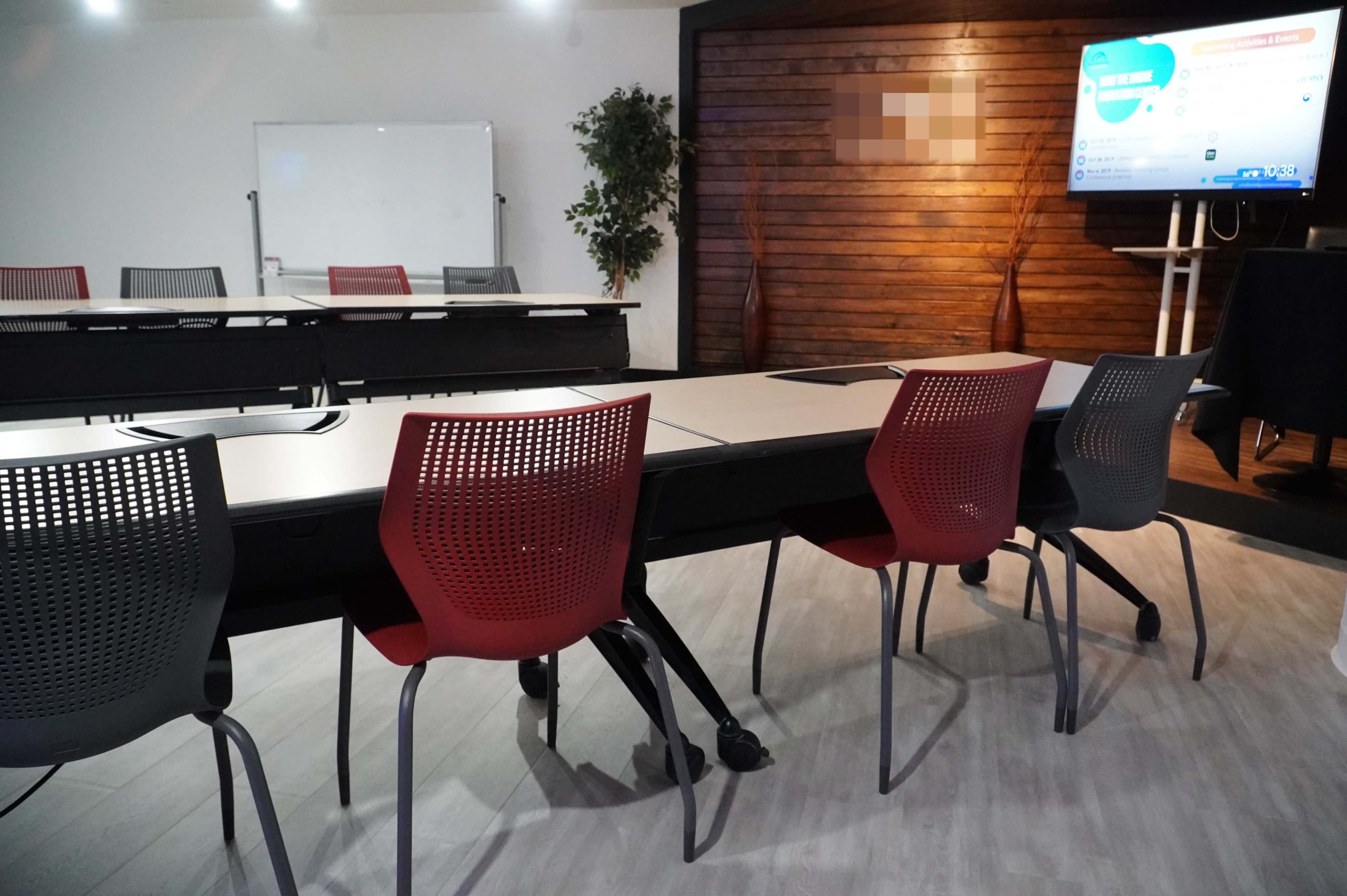 A modern meeting room with several tables and chairs arranged, a large screen displaying information, and a whiteboard in the background.