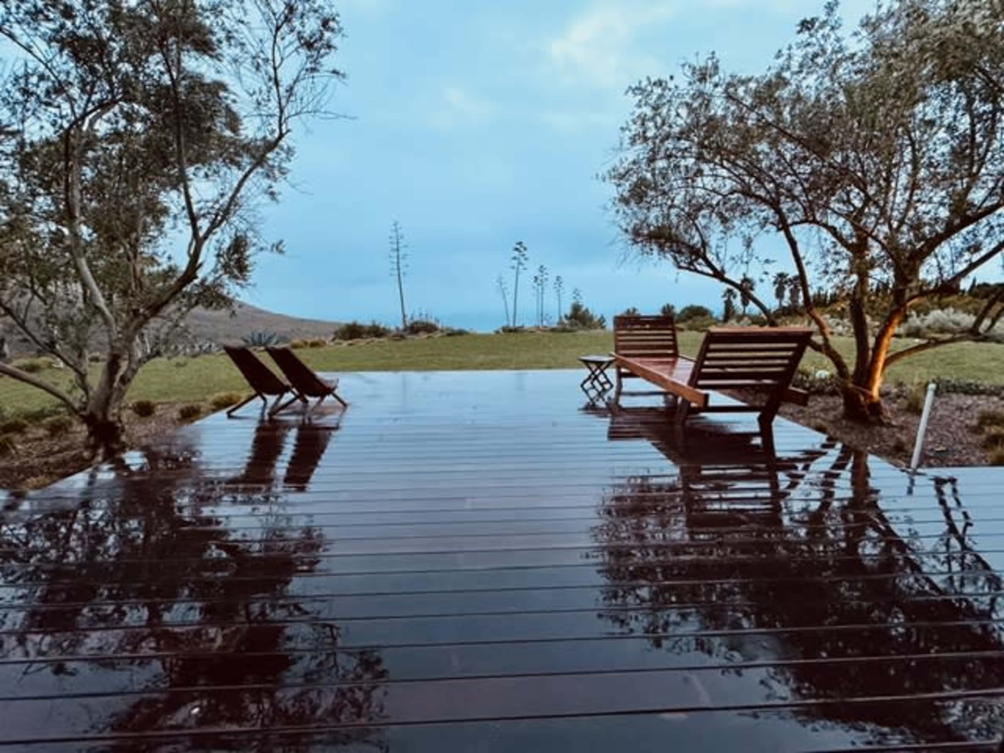 A wooden deck is visible after rain, featuring two lounge chairs and surrounded by trees and grass under an overcast sky.
