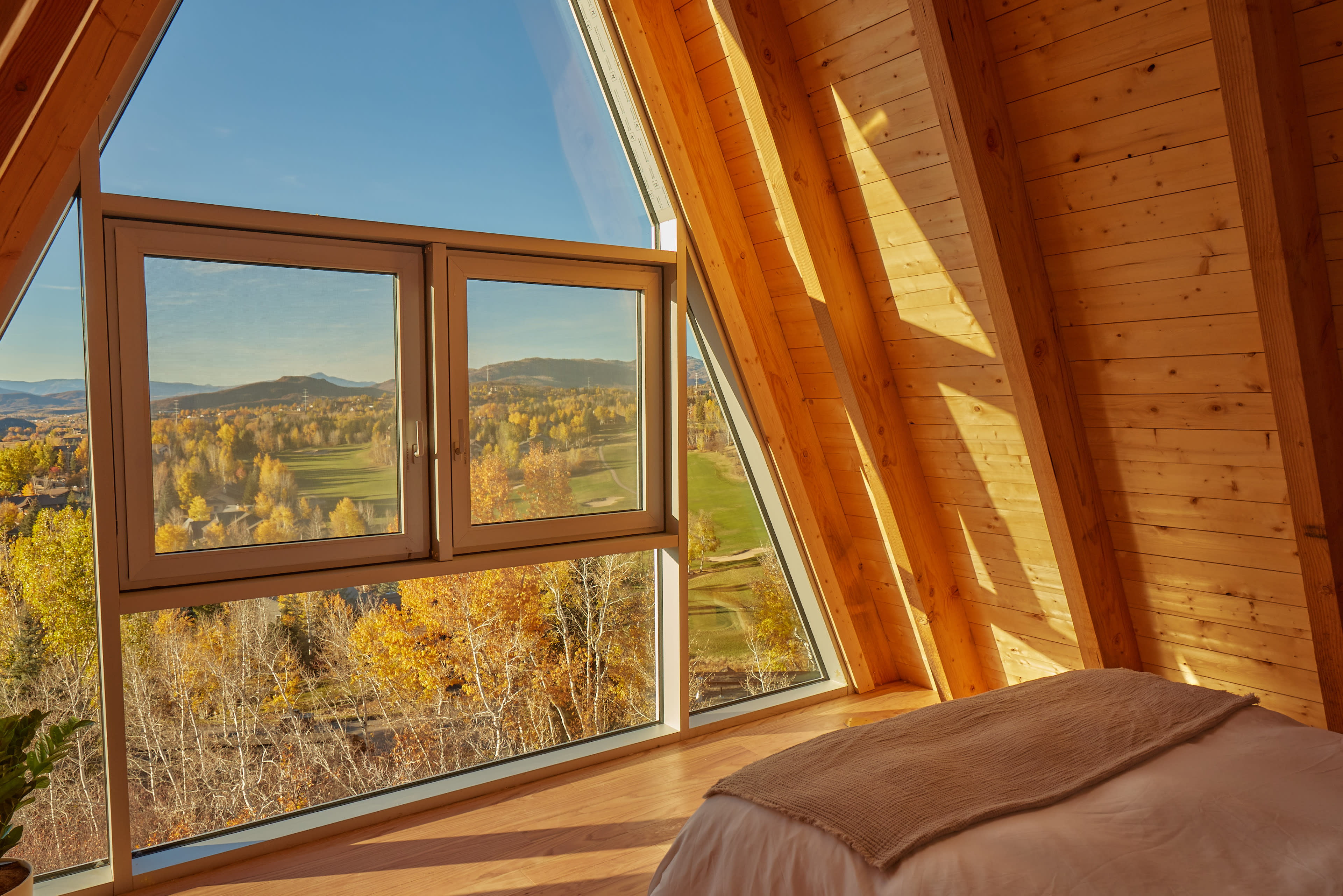 The image shows a wooden interior room with a large angled window offering a view of autumn-colored trees and rolling hills.