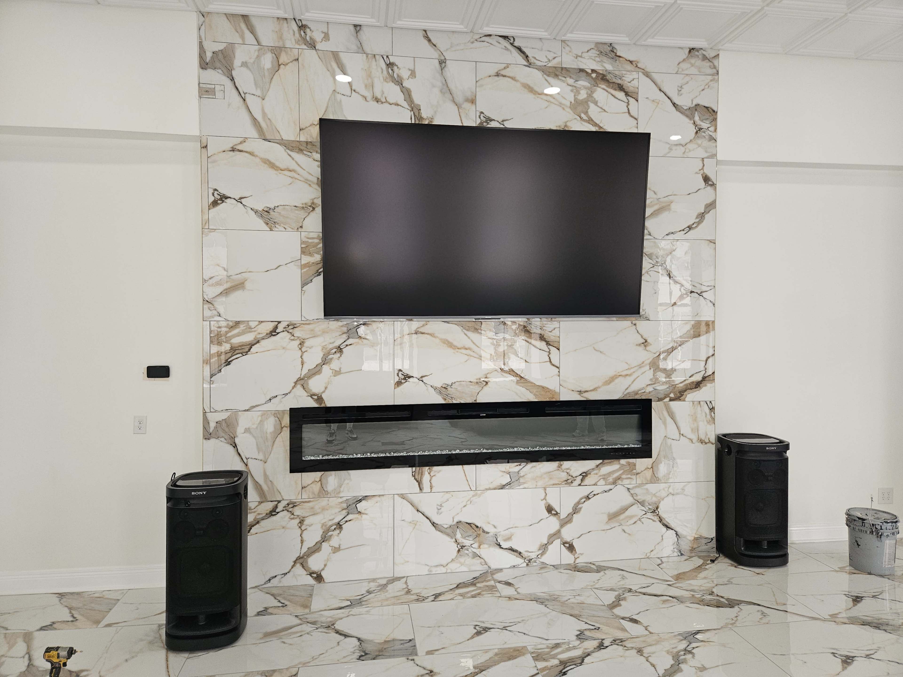 A large flat-screen television is mounted on a marble accent wall, with a modern electric fireplace below it and two speakers on either side.