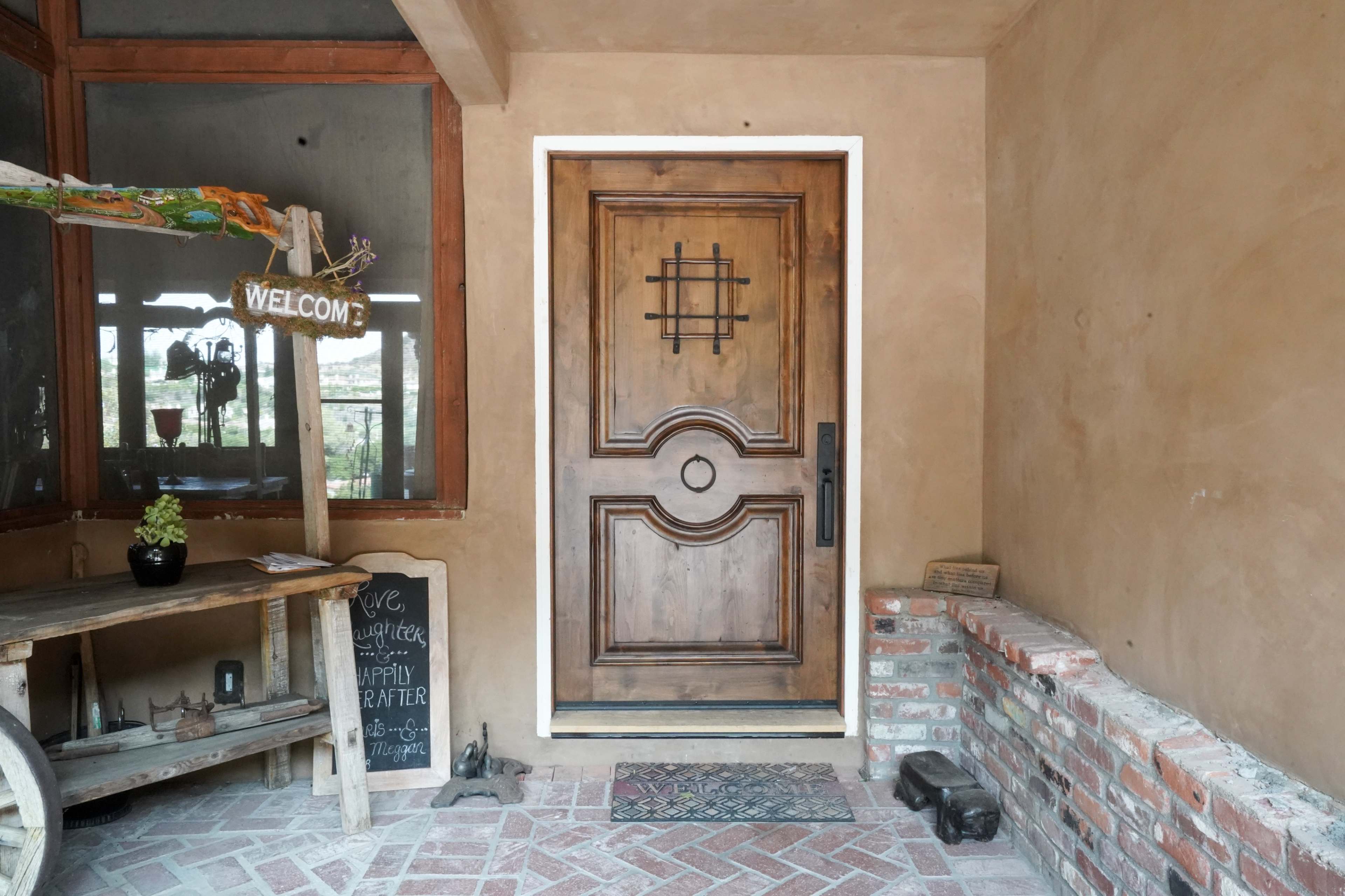 The image shows a wooden front door with a decorative grid pattern, surrounded by a rustic entrance featuring a welcome sign and a small table with a chalkboard.