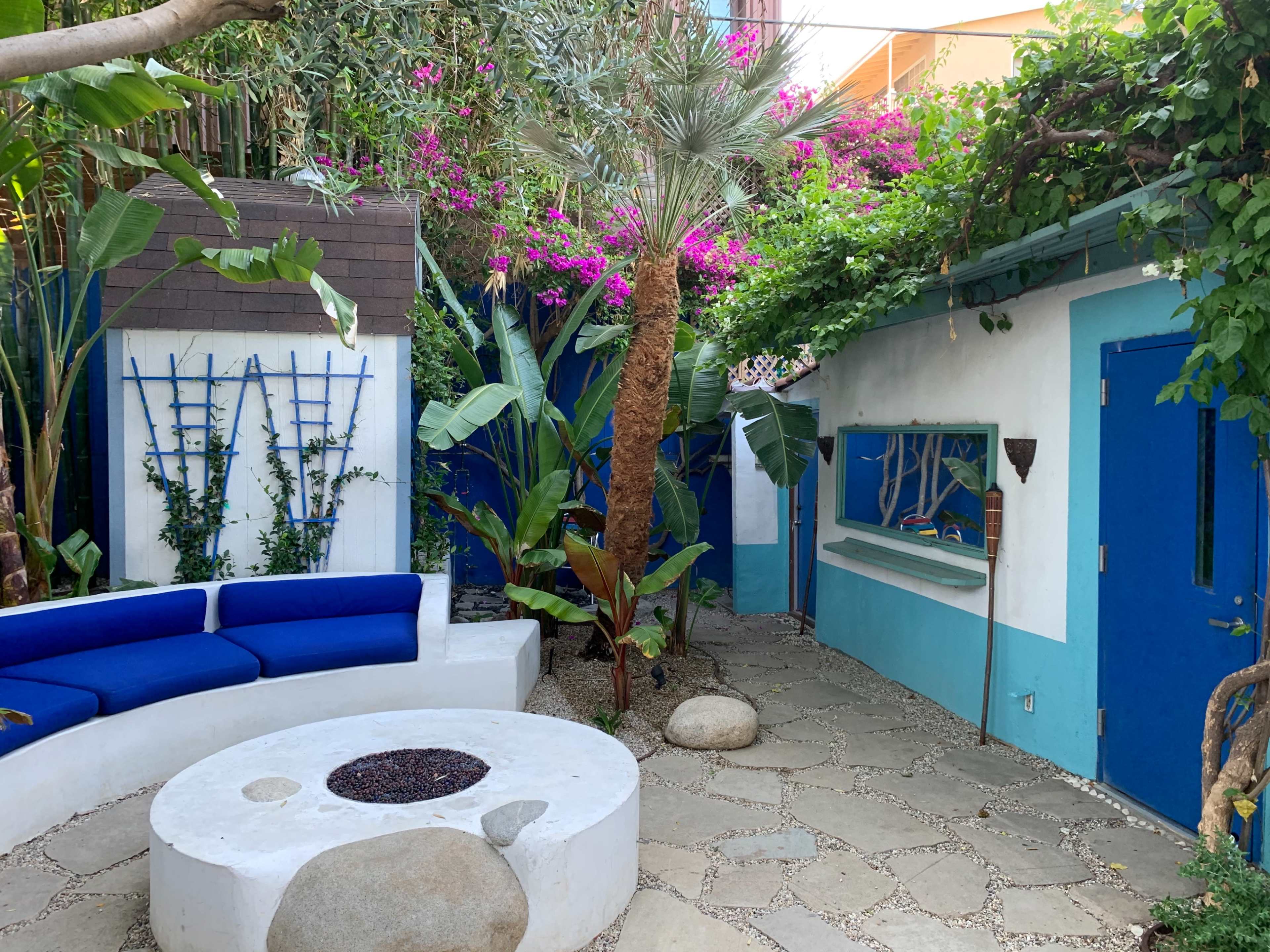 A courtyard features a circular fire pit surrounded by a blue sofa, flanked by tropical plants and vibrant flowers against a white and blue wall.