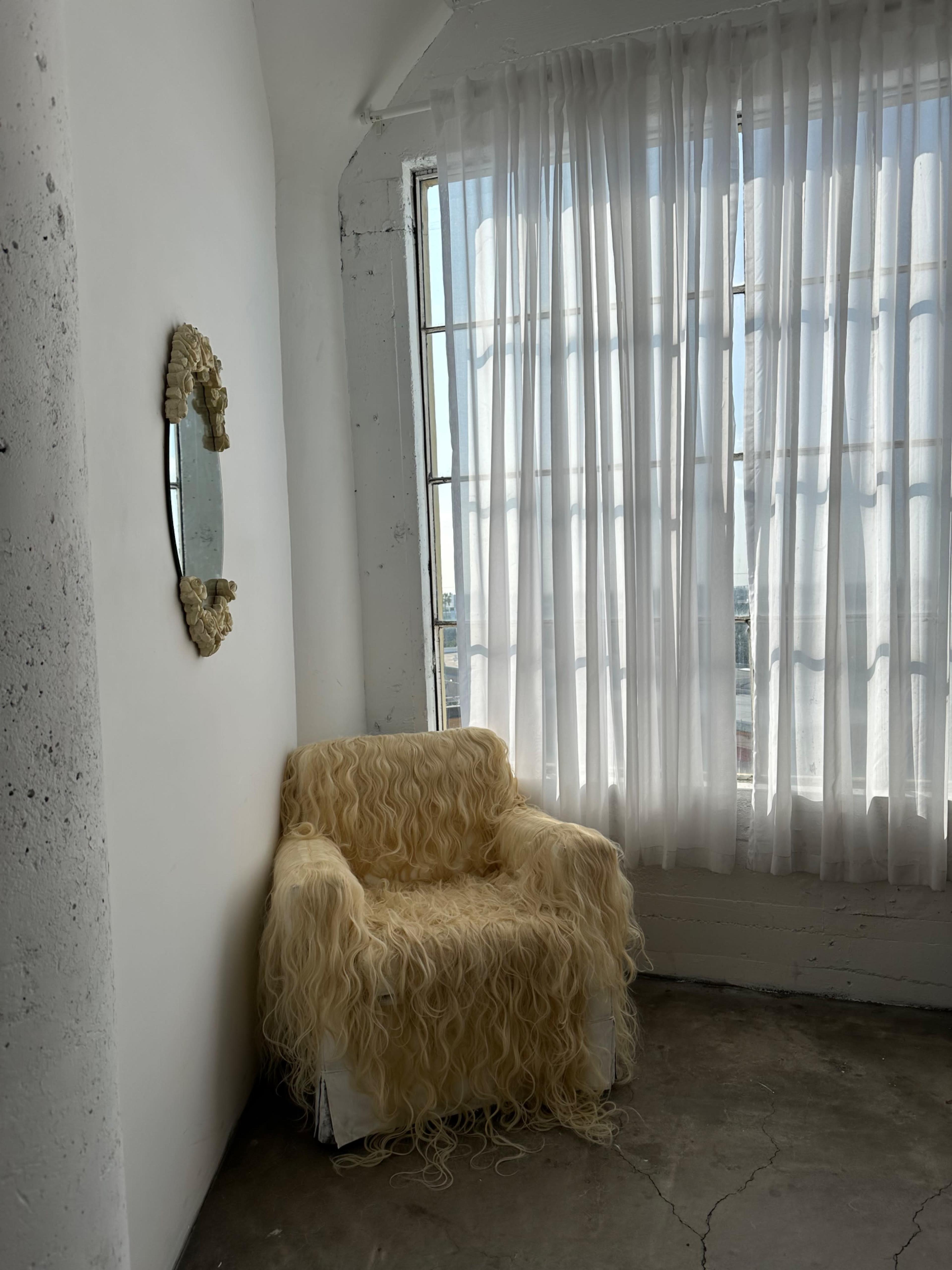 A textured chair draped in long, light-colored fibers sits beside a window with sheer white curtains in a minimalist room.