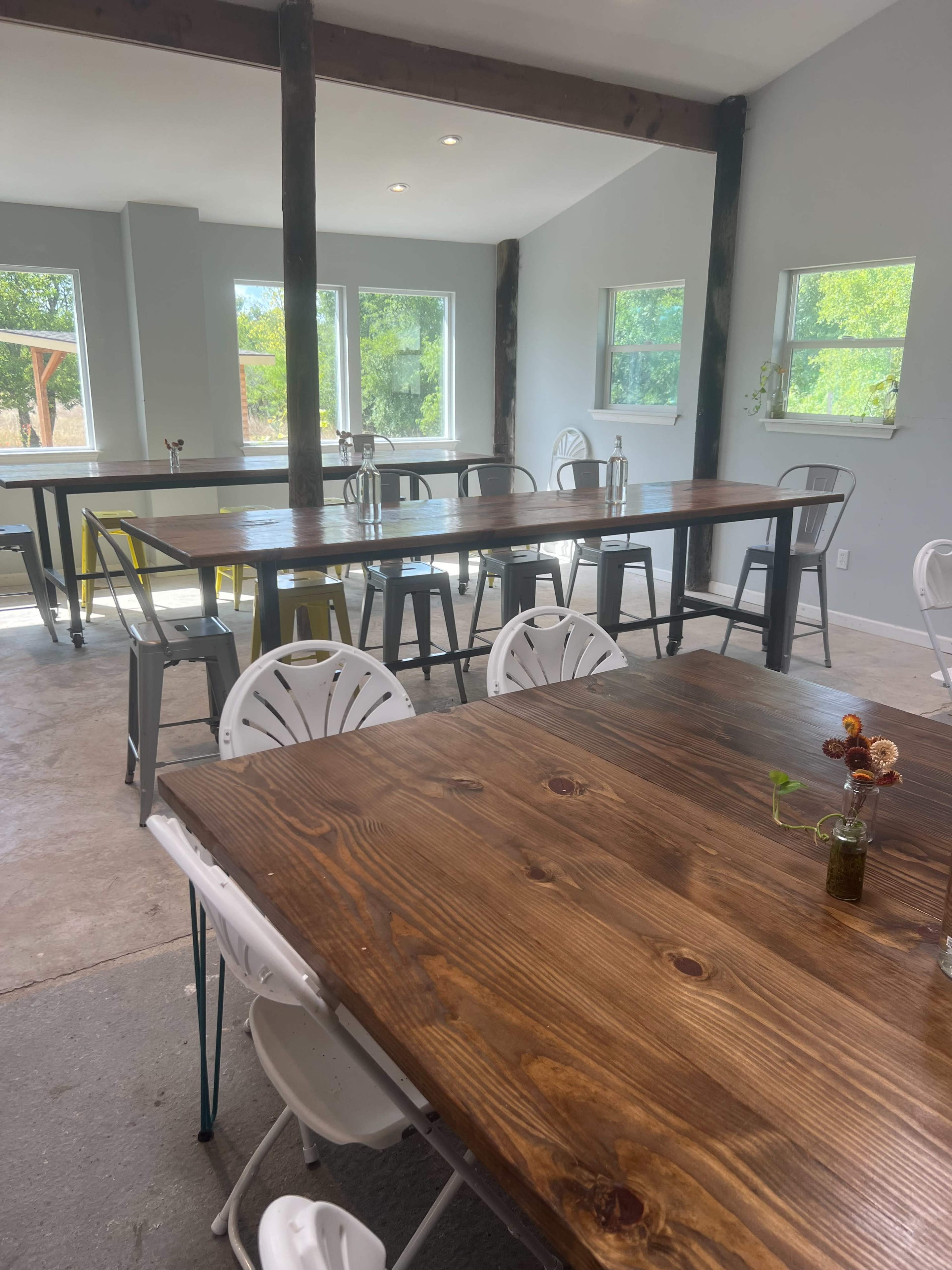 A spacious interior with several long wooden tables and white chairs, illuminated by natural light from multiple windows.