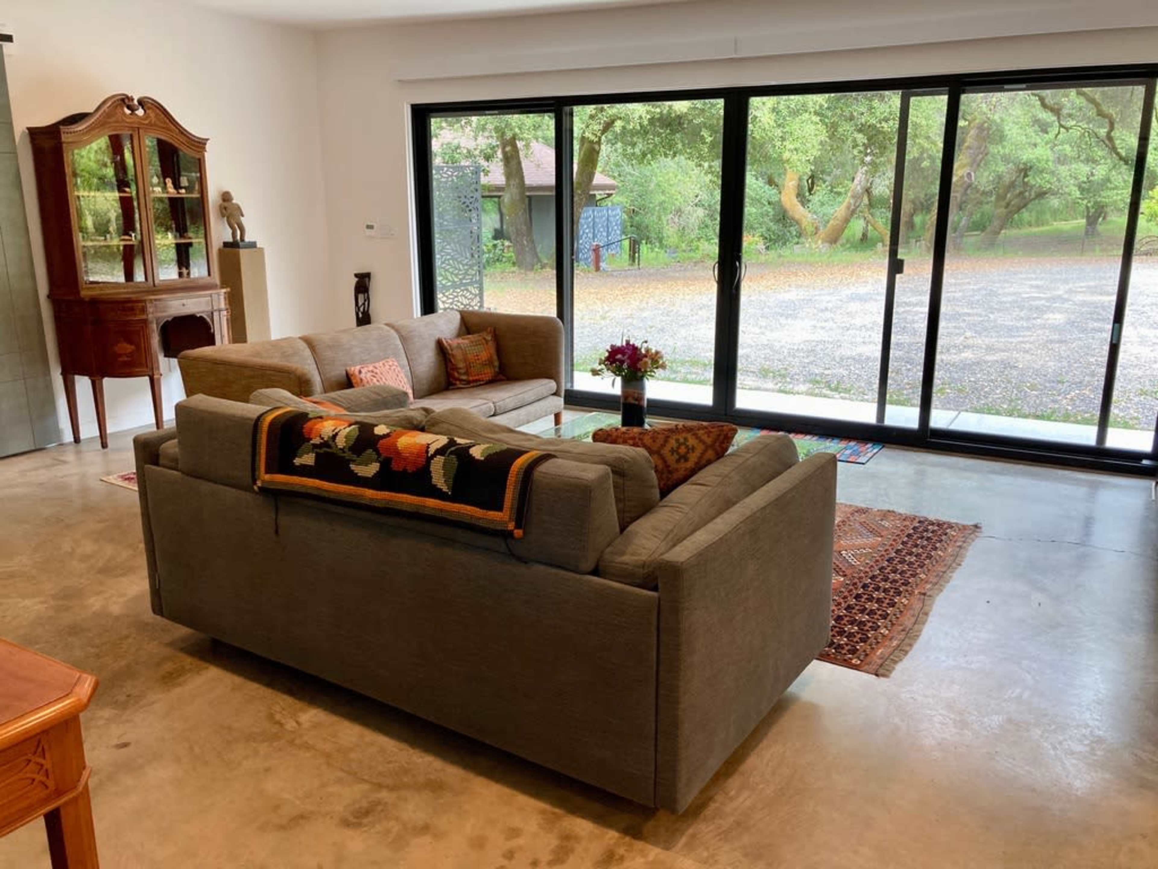 A gray sofa is positioned in a living room with large sliding glass doors that open to a view of trees and a gravel pathway outside.