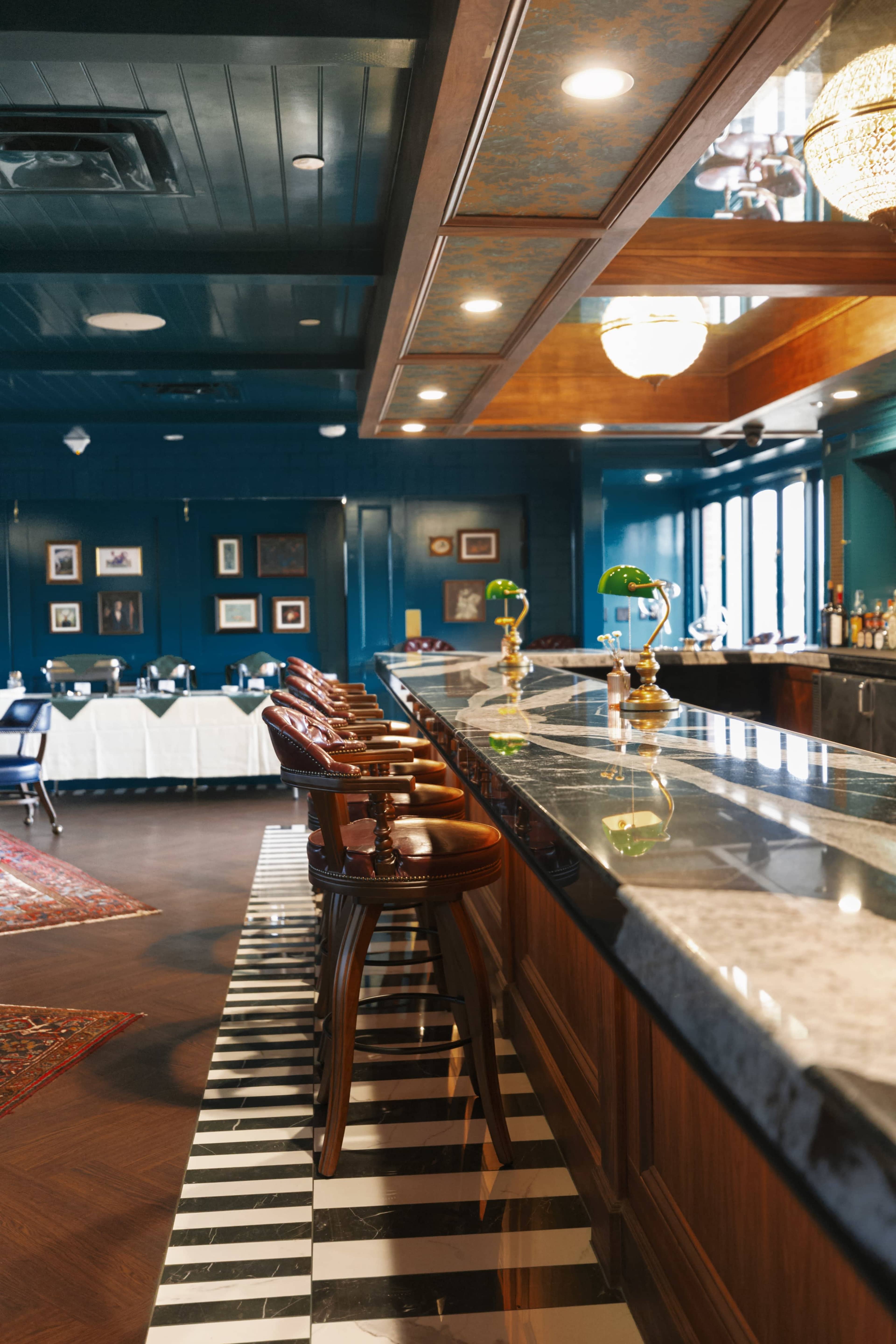 The image shows a stylish bar with wooden stools and a marble countertop, featuring a checkerboard floor pattern and a cozy dining area in the background.