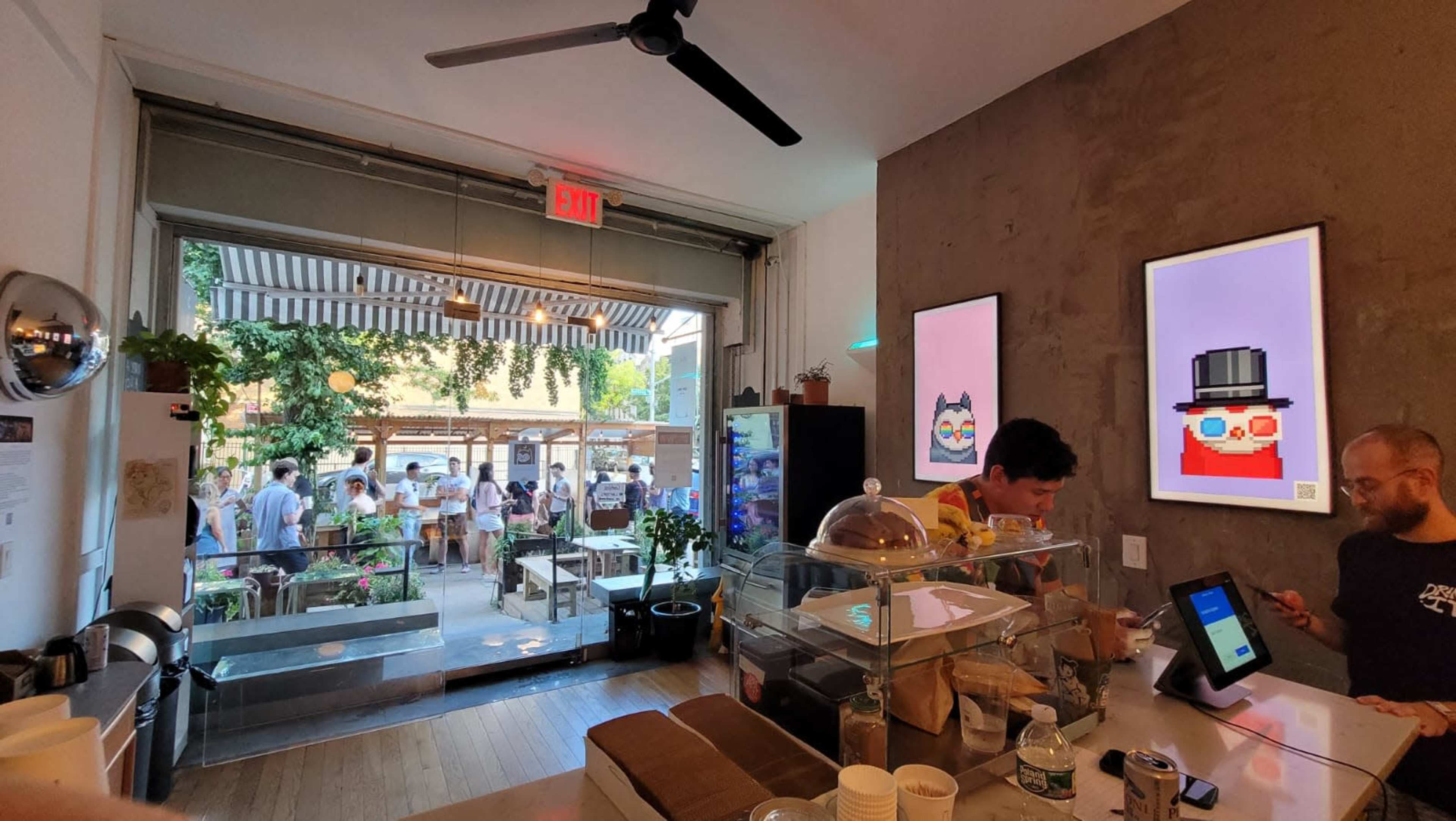 The scene shows the interior of a cafe with a counter, two colorful framed artworks on the wall, and customers in the outdoor area visible through an open entrance.