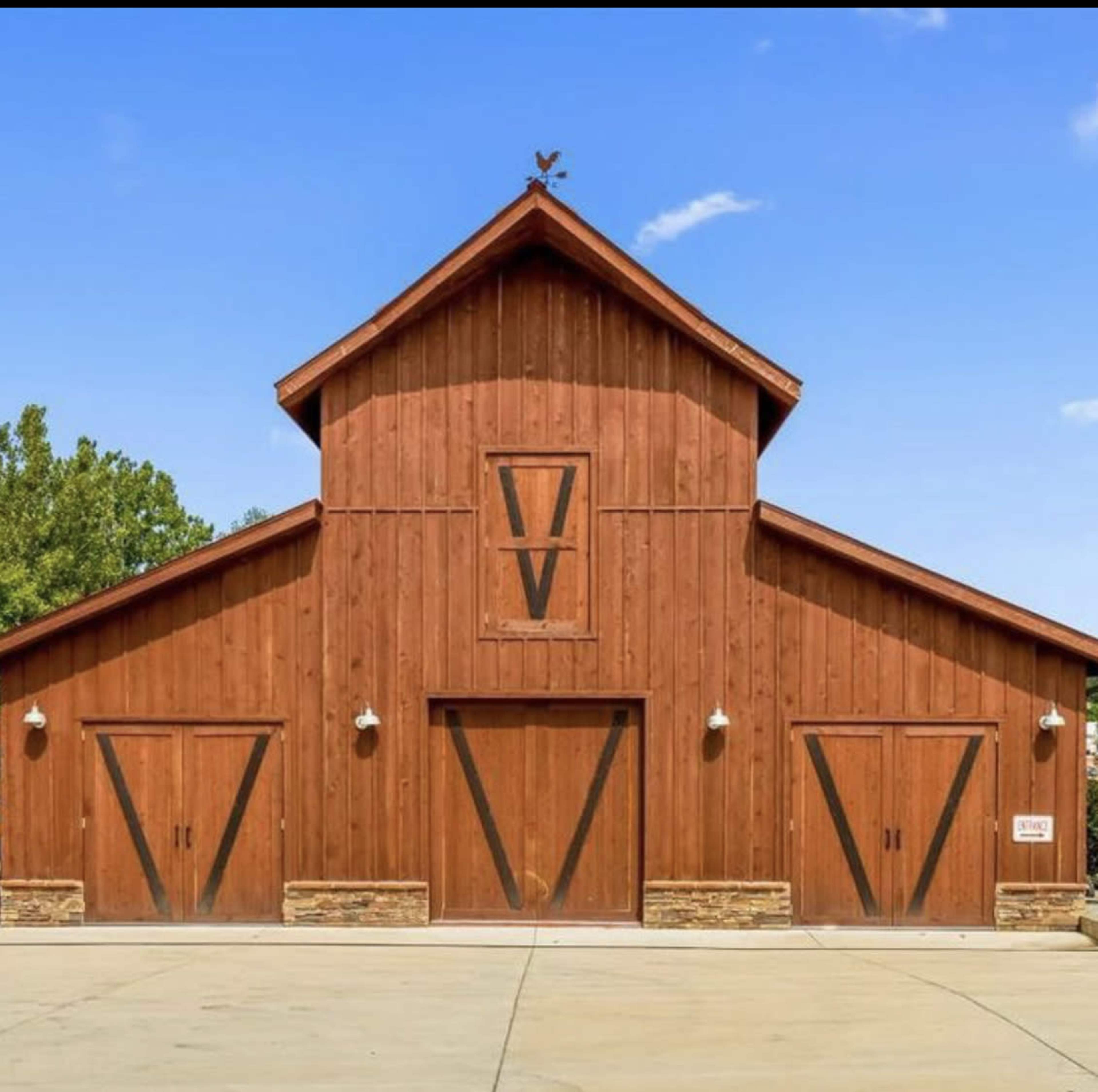 The image shows a large wooden barn with a steeply pitched roof, featuring two large double doors and a weather vane on top.