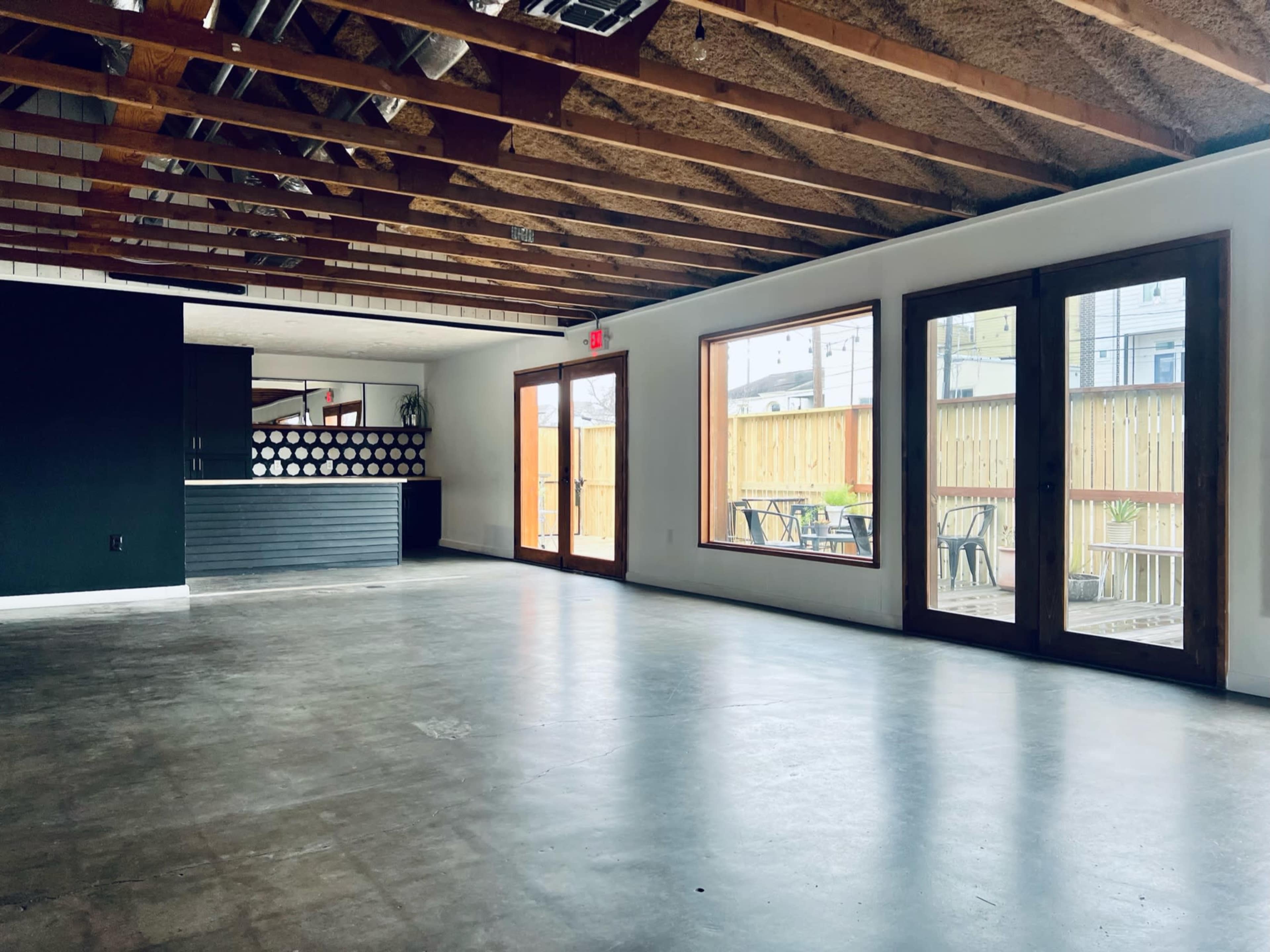 An empty room with exposed beams, concrete flooring, large windows, and a counter area in the background.