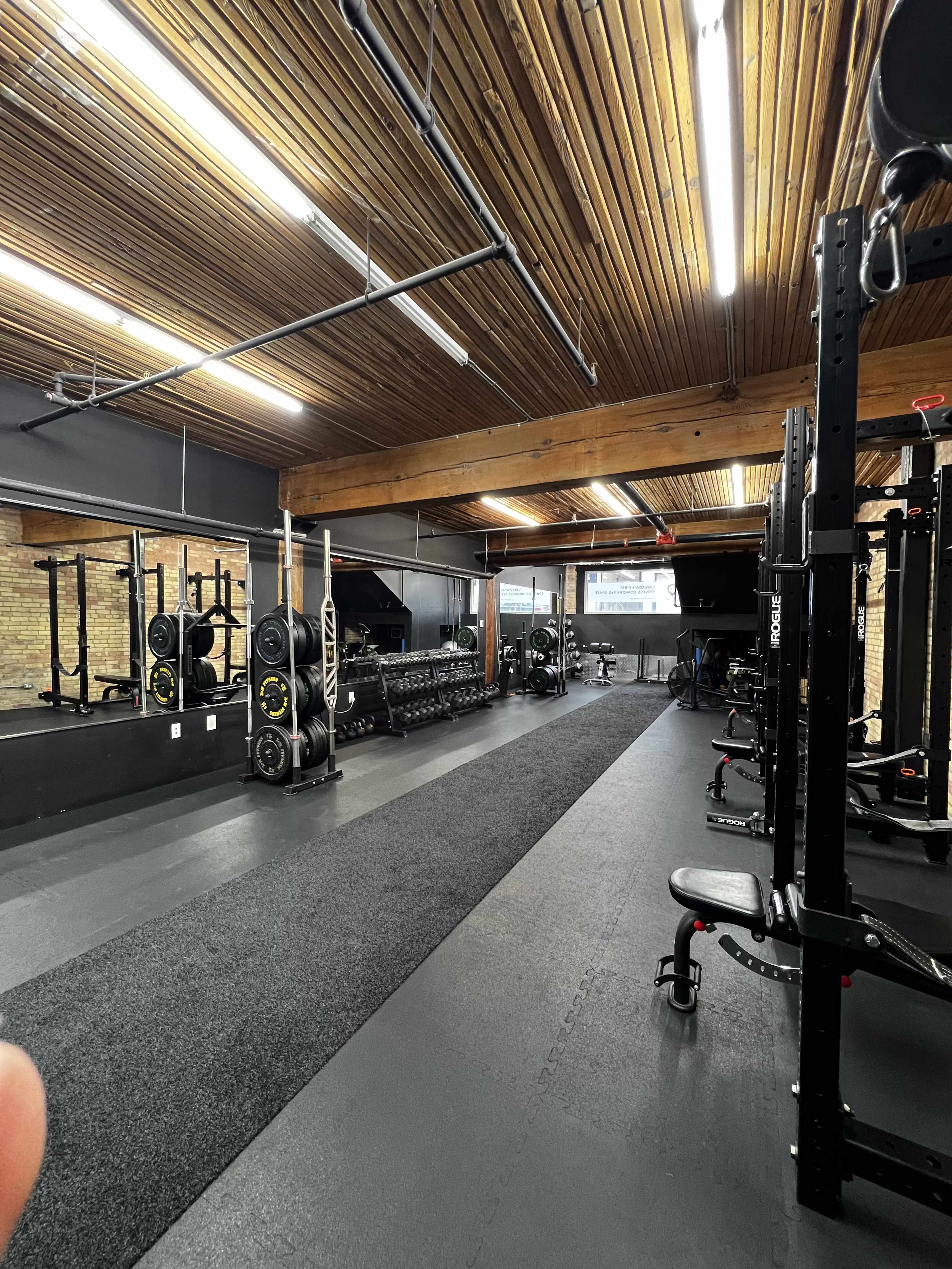 An empty gym with weightlifting equipment, benches, and a rubber floor, illuminated by overhead lights and featuring wooden ceiling beams.