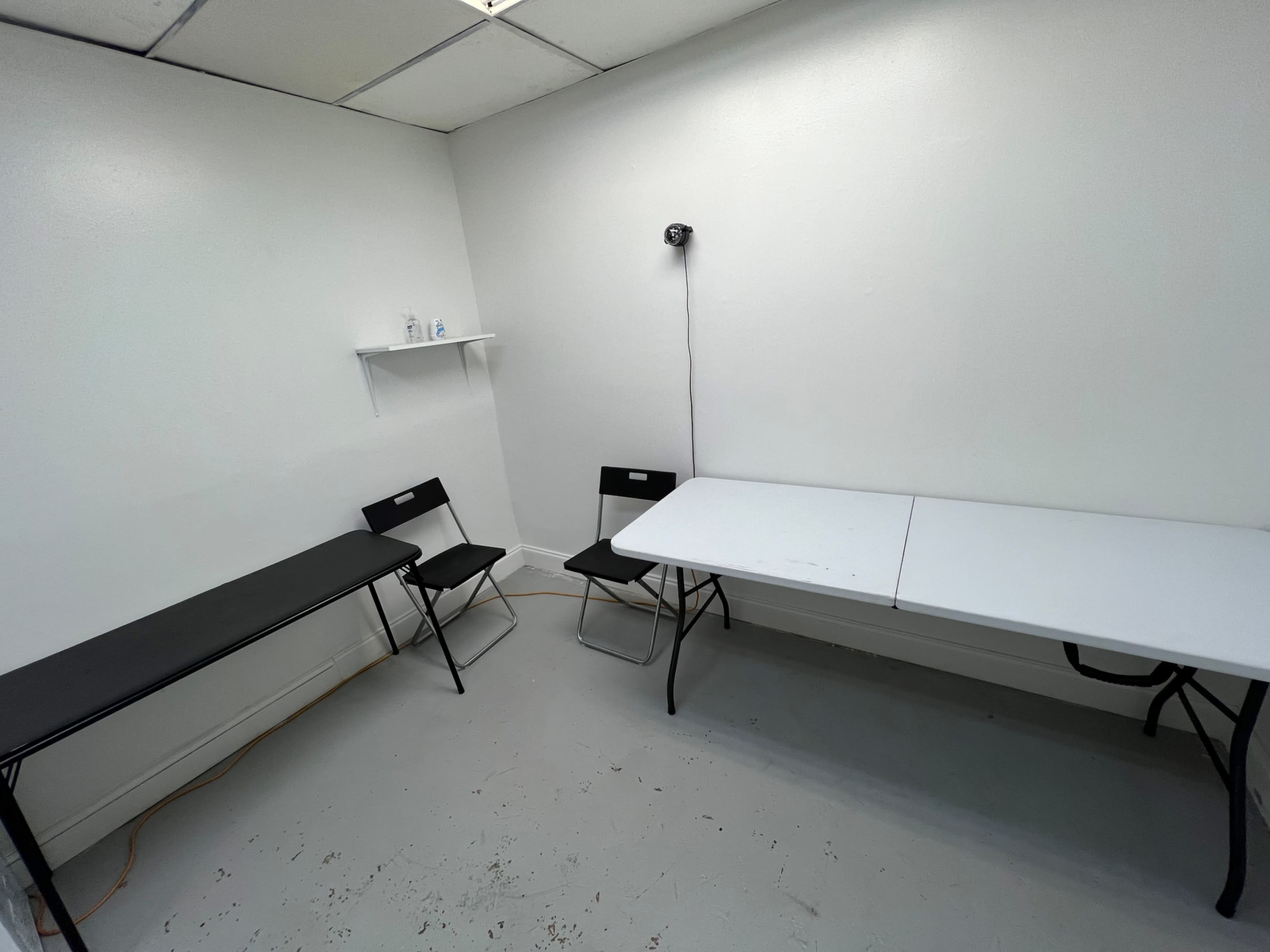 A small, empty room with bare white walls, a light-colored floor, two folding tables, and two black folding chairs.