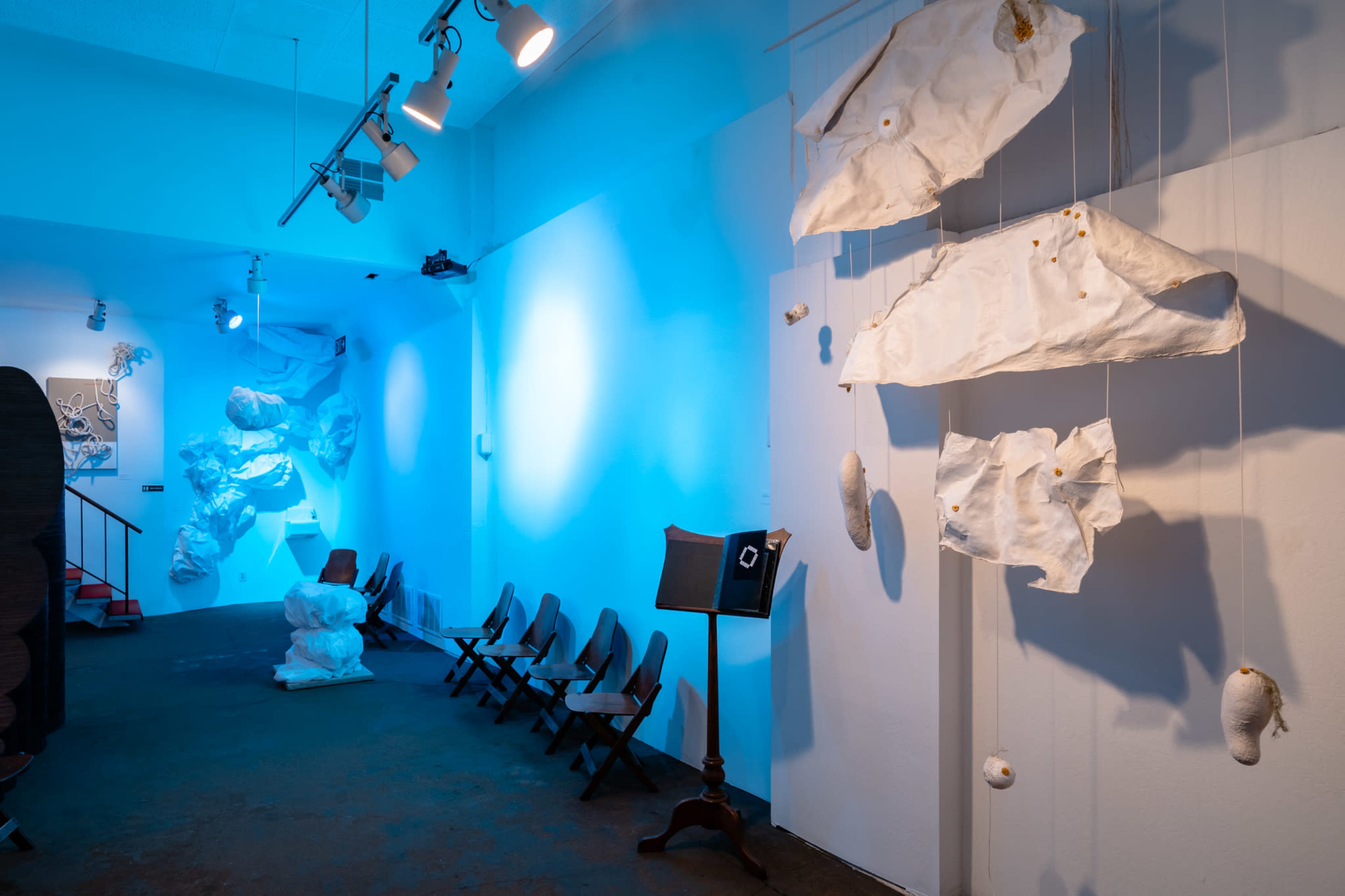 An art gallery features white sculptural forms hanging from the ceiling, illuminated by blue lighting, with wooden seating arranged along one side.
