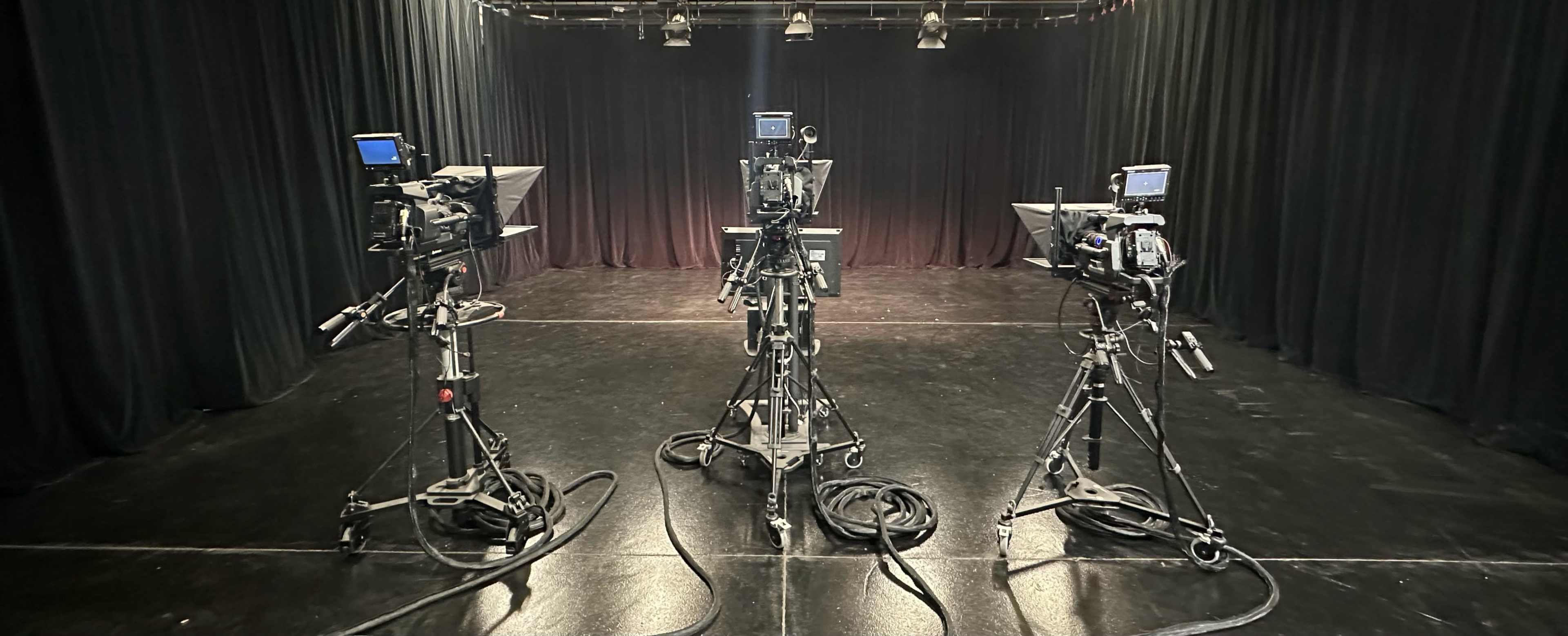 Three camera setups on tripods are positioned on a dark stage with black curtains in the background.