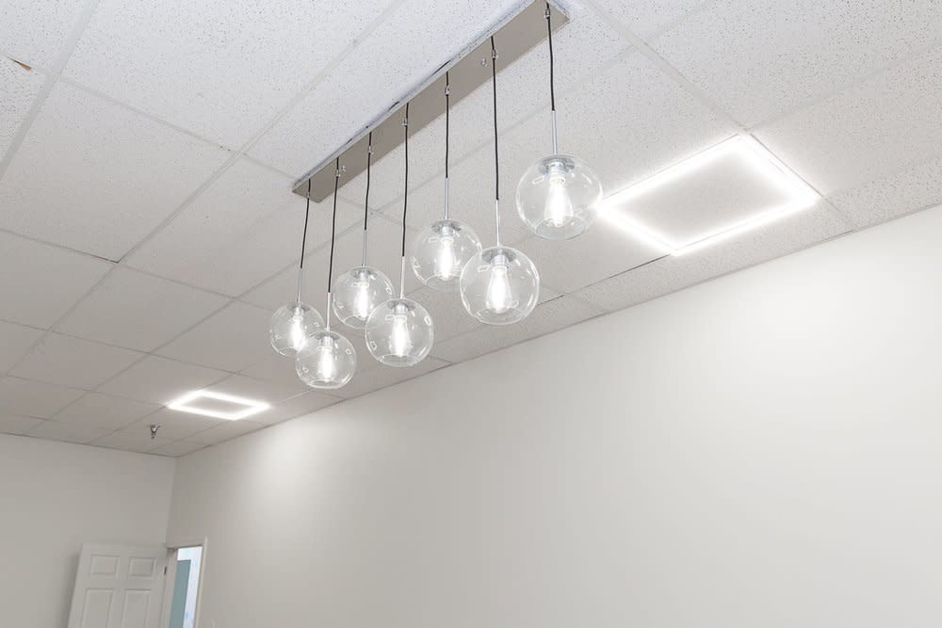 A modern lighting fixture with seven glass pendant lights hanging from a rectangular ceiling mount in a room with a white, minimalistic design.
