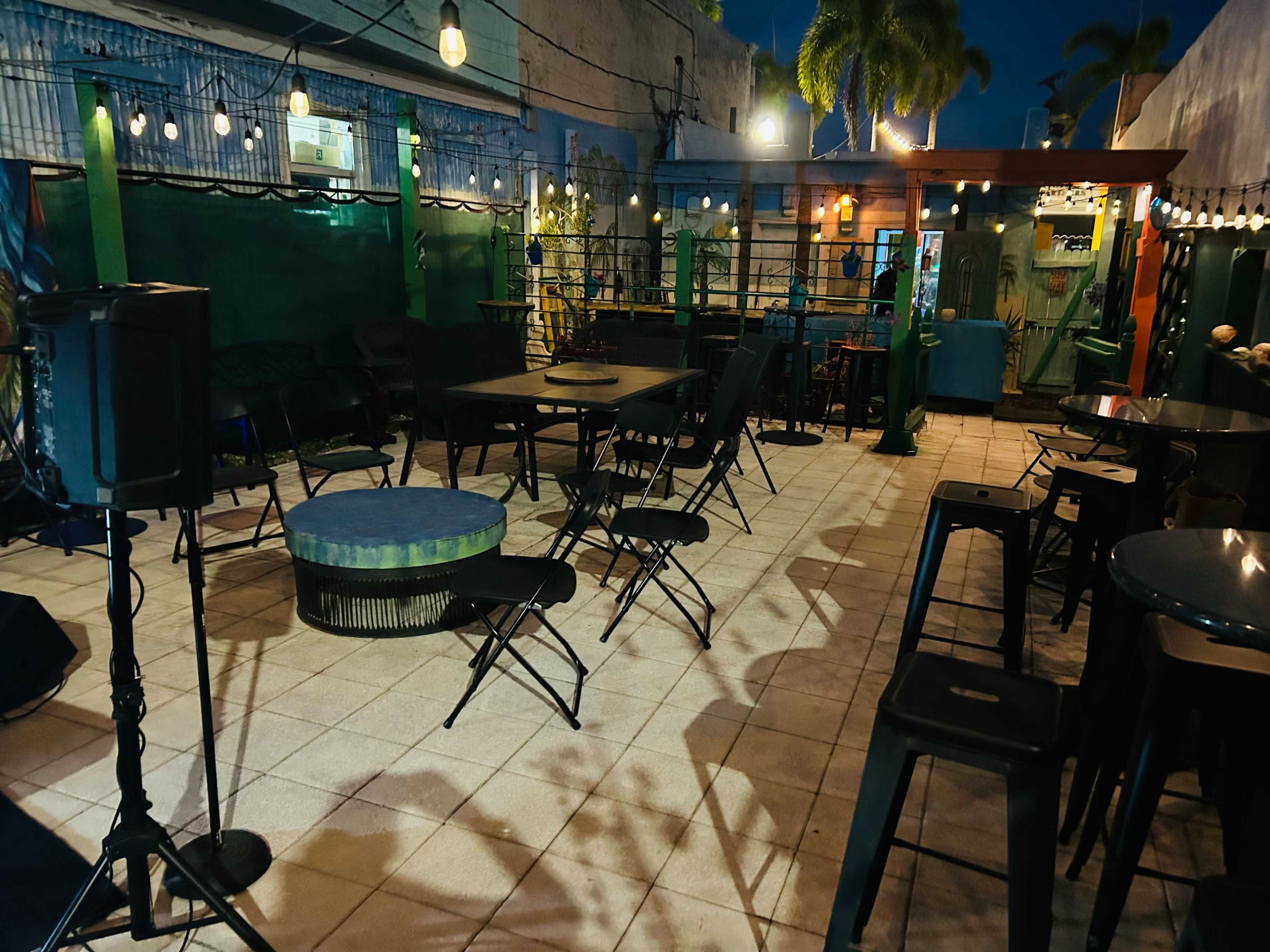 The image shows an outdoor seating area of a bar featuring black tables and chairs, string lights overhead, and a partially enclosed space with plants in the background.