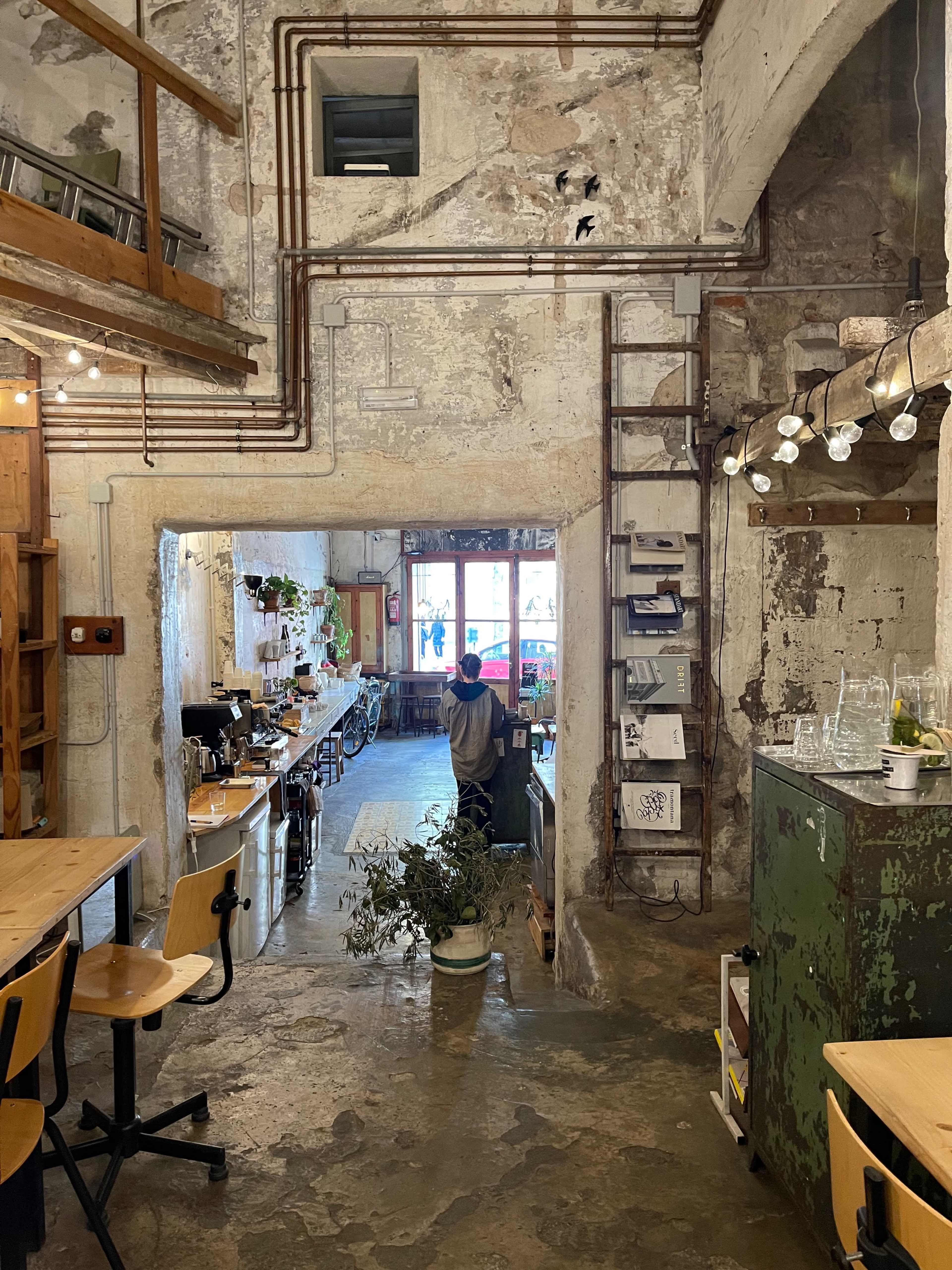 A person stands in a rustic café with exposed brick walls, visible plumbing, and wooden tables arranged for seating.