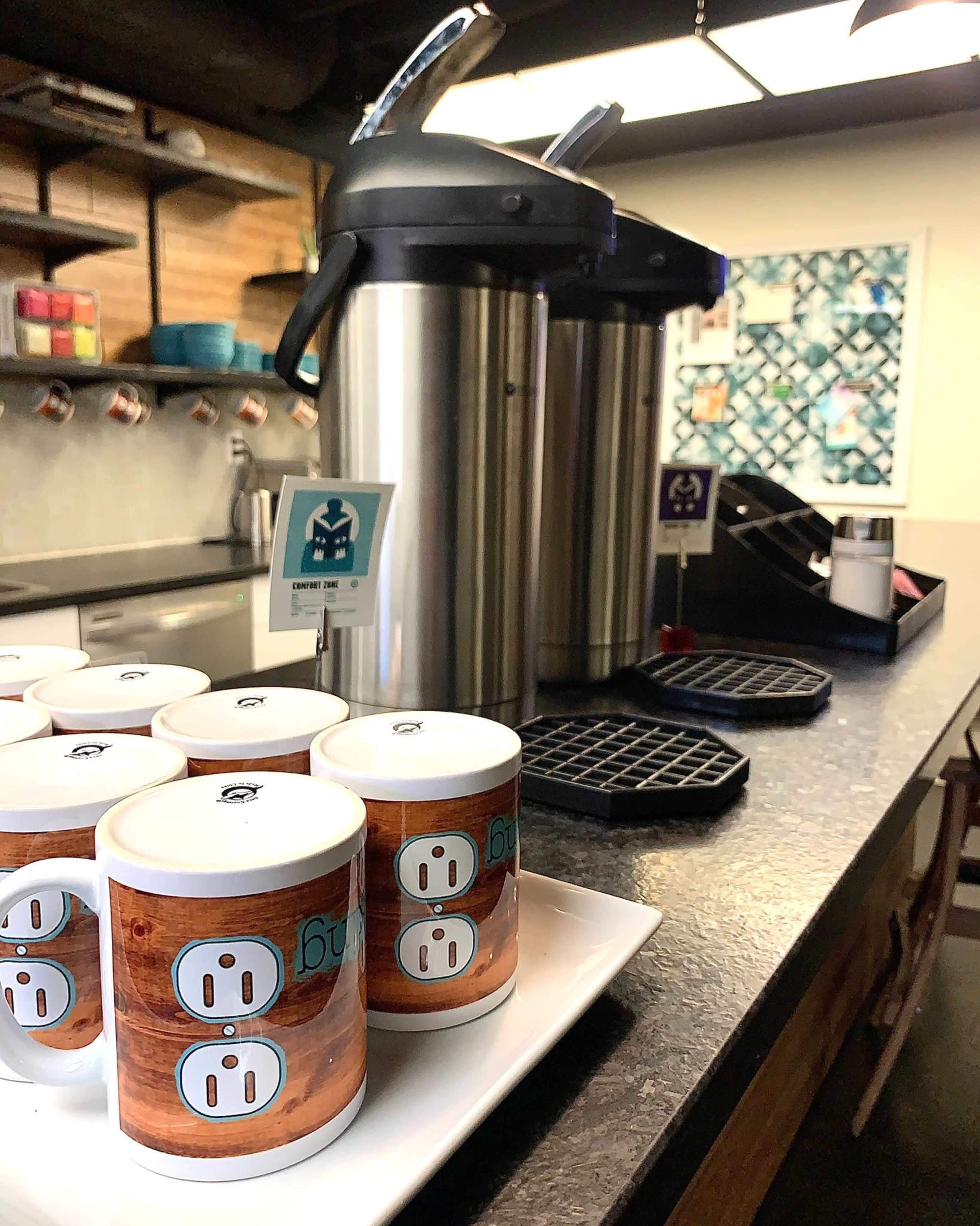 A countertop with two large beverage dispensers and several mugs arranged on a tray, alongside a waffle maker and wall displaying artwork.