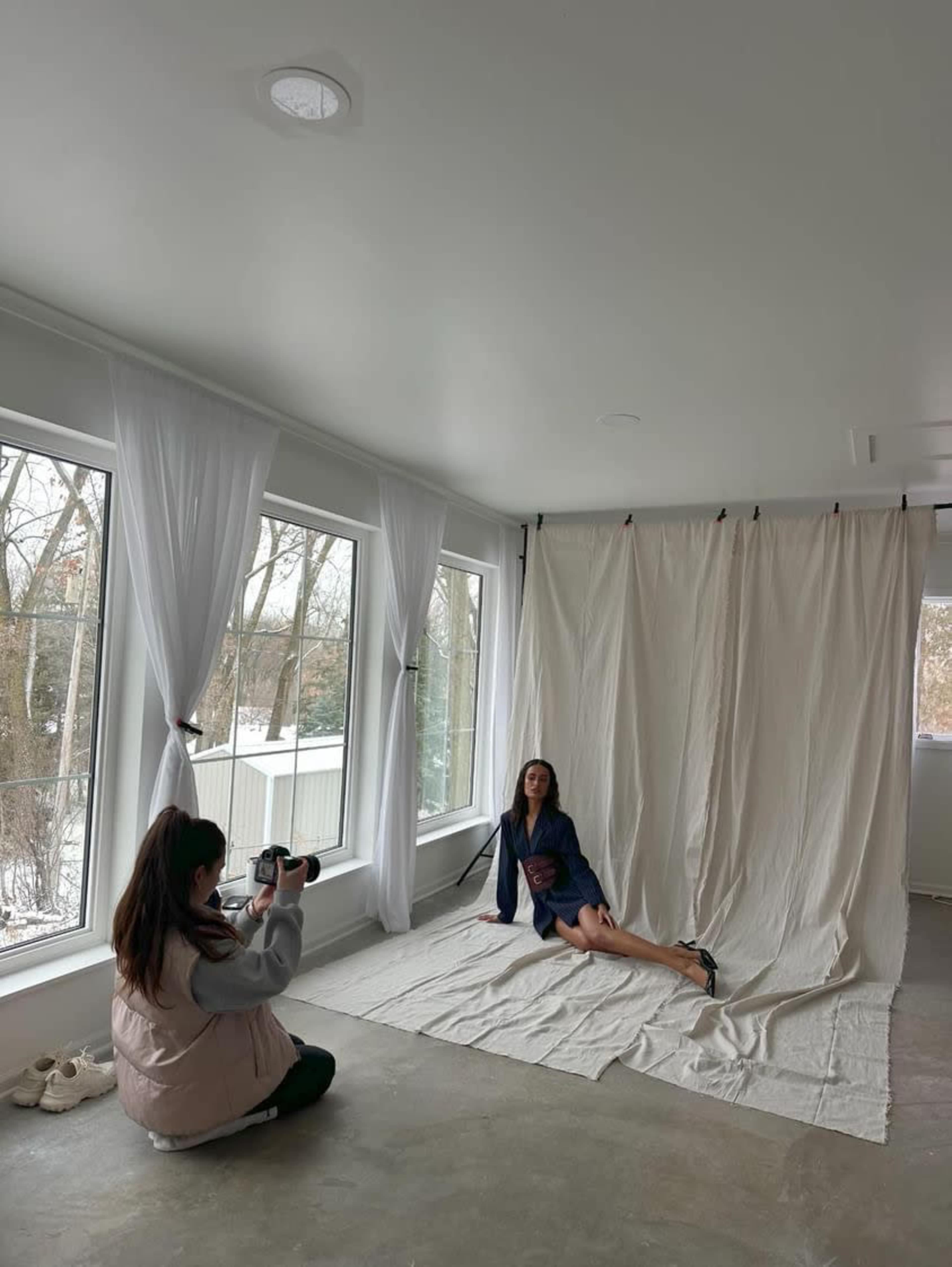 A photographer captures a model posing on a light-colored backdrop in a spacious room with large windows.