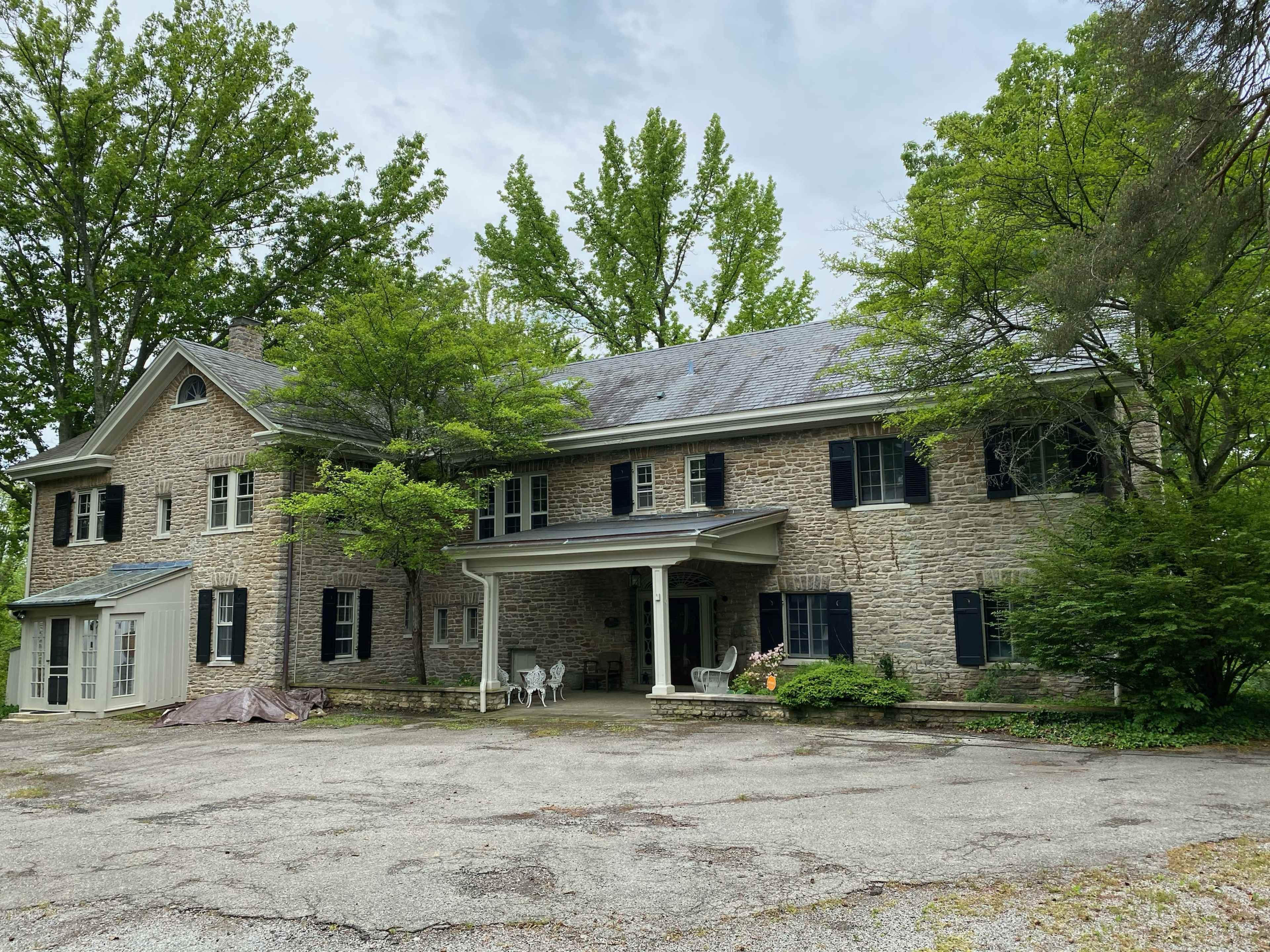 A large stone house surrounded by trees, with a gravel driveway and a covered porch in front.