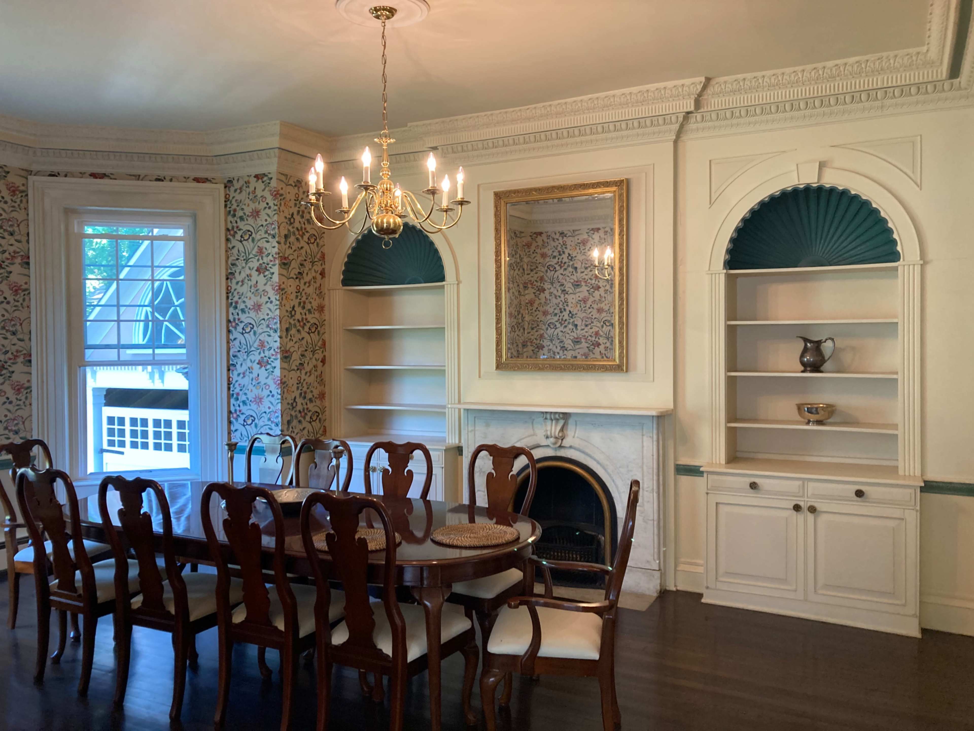 A dining room with a long wooden table surrounded by chairs, a chandelier overhead, and built-in shelves beside a decorative fireplace.