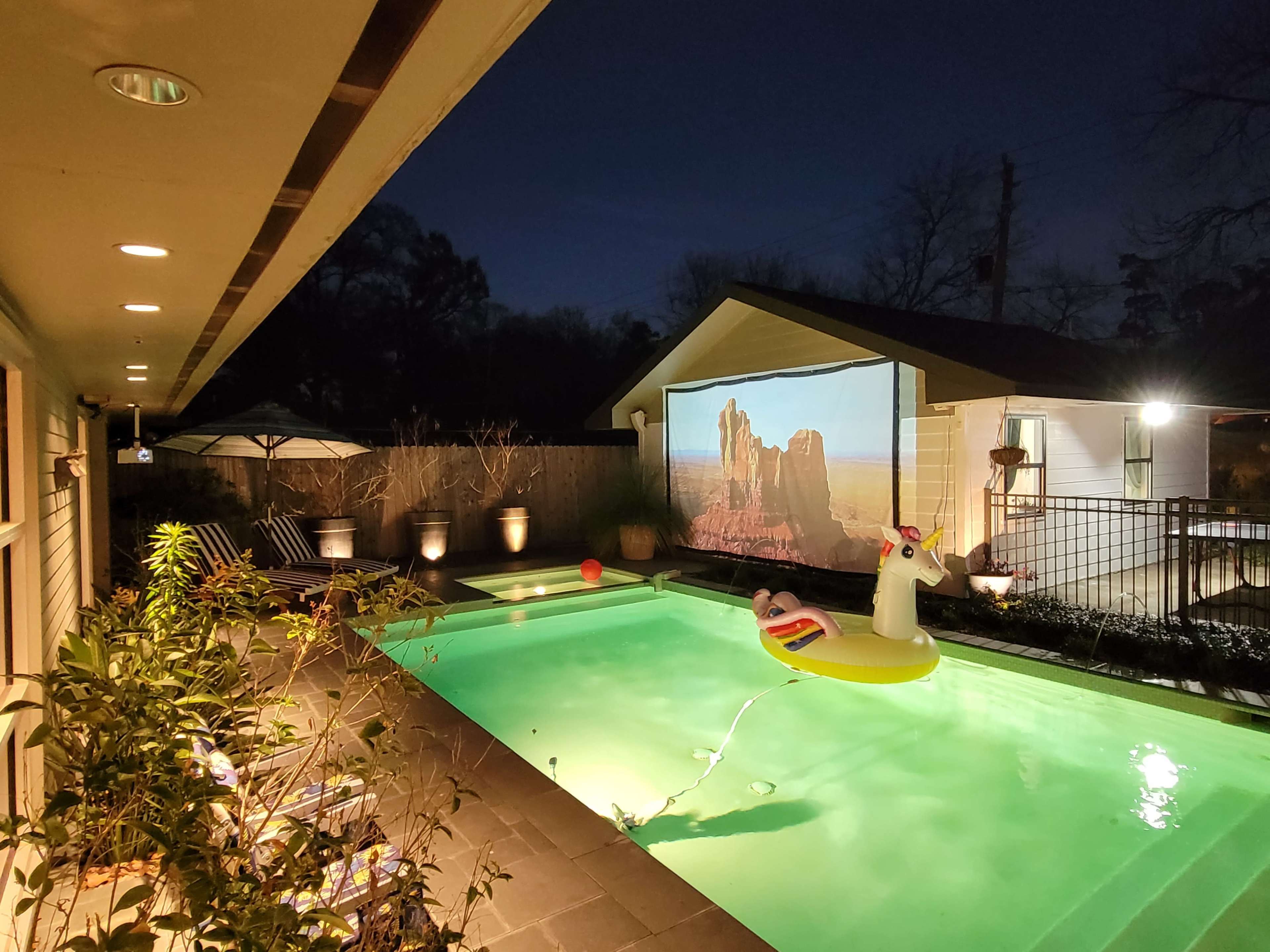 A pool area features a unicorn float, a movie screen showing a landscape, and illuminated outdoor plants under a night sky.