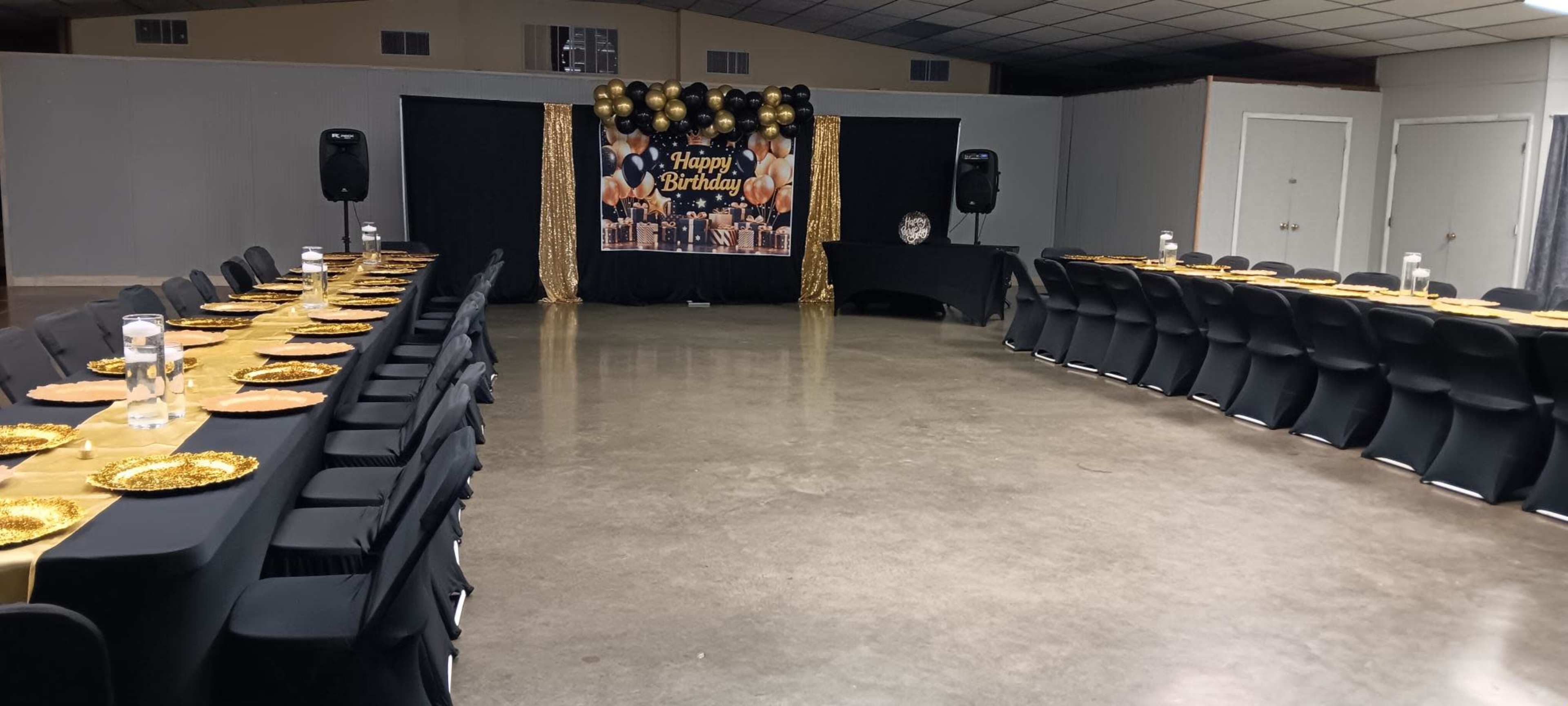 The image shows a large indoor event space set up for a birthday celebration, featuring rows of tables with black tablecloths, gold accents, and a decorated backdrop with balloons and a "Happy Birthday" sign.