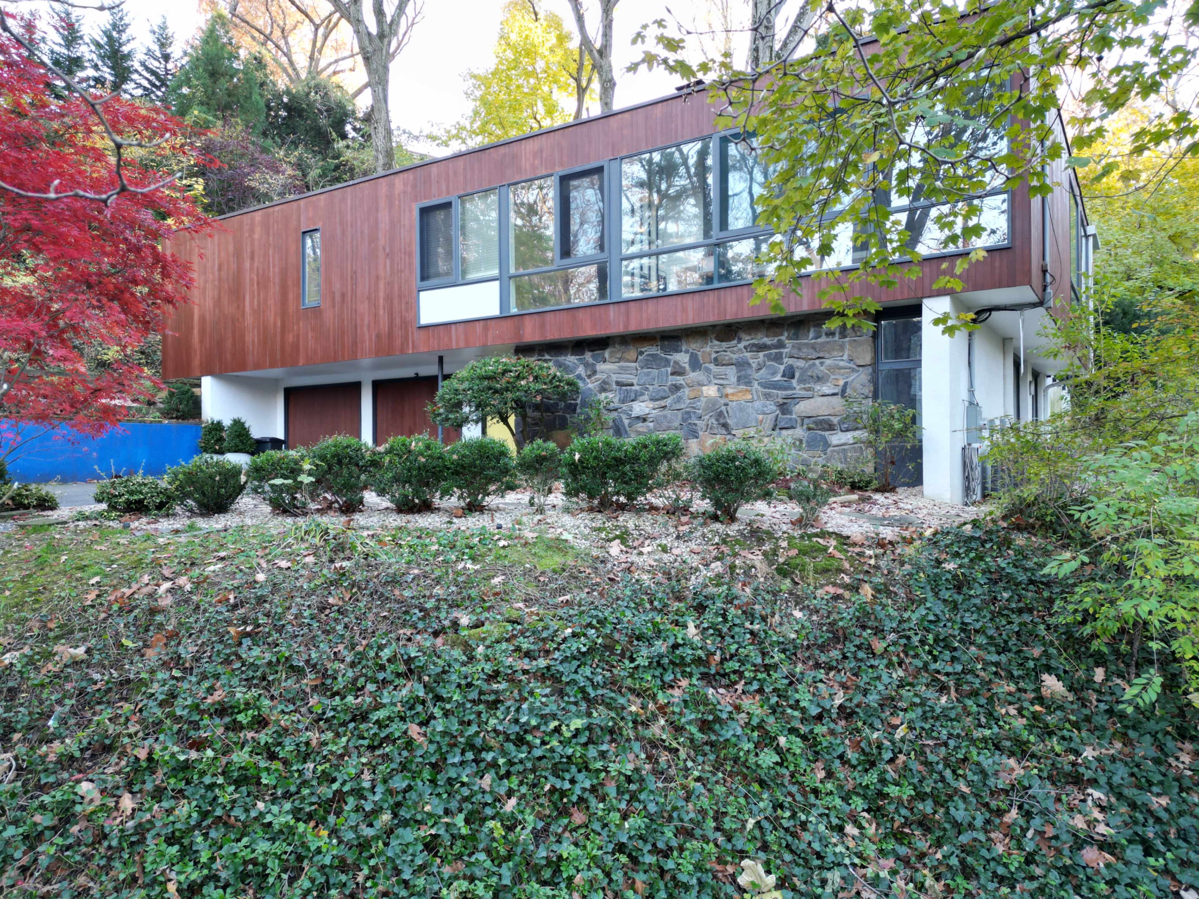 The image shows a modern house with a wooden and stone facade, situated on a sloped landscape surrounded by greenery and trees.