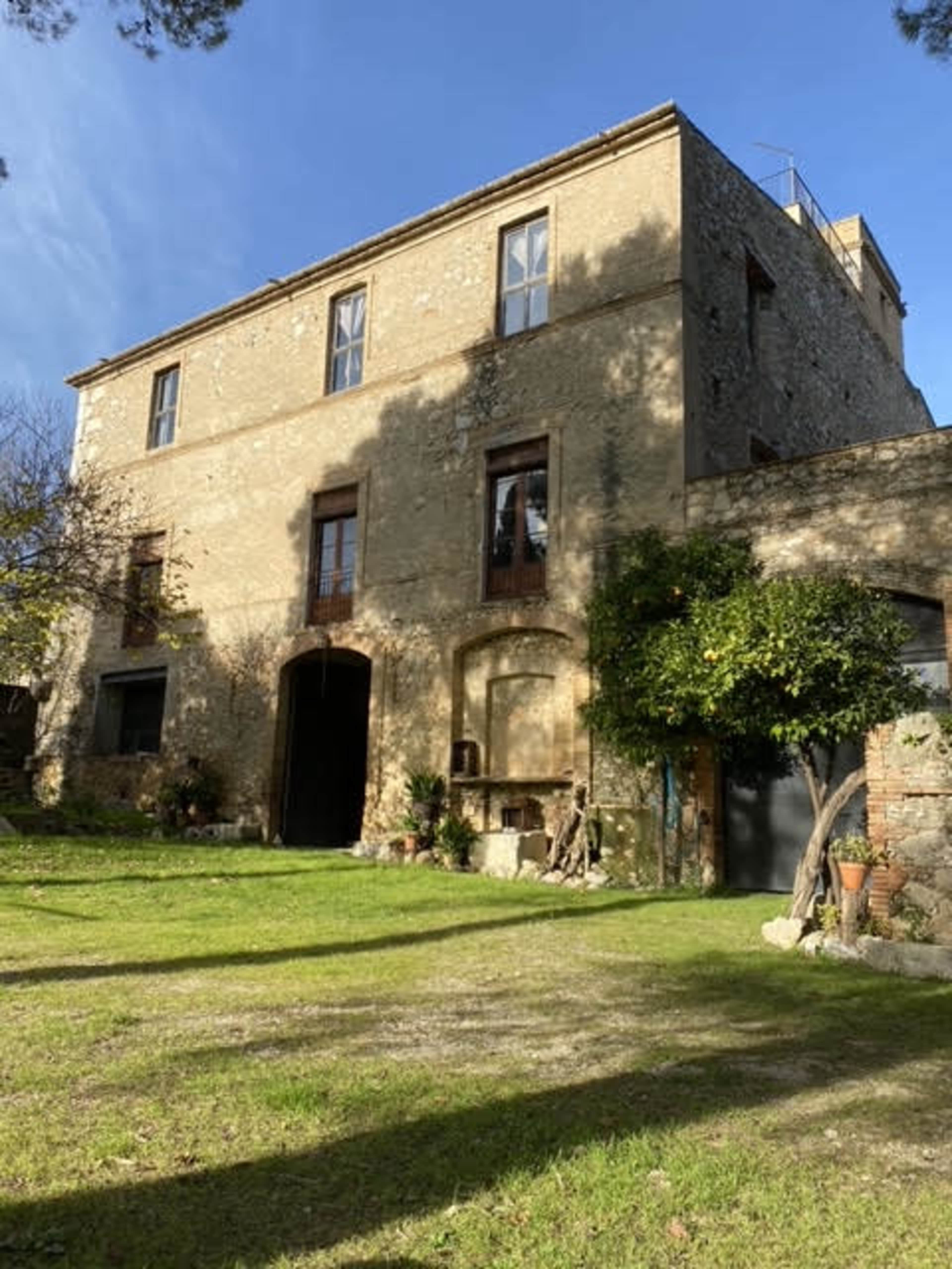 A three-story stone building with rectangular windows stands surrounded by a grassy area and trees.