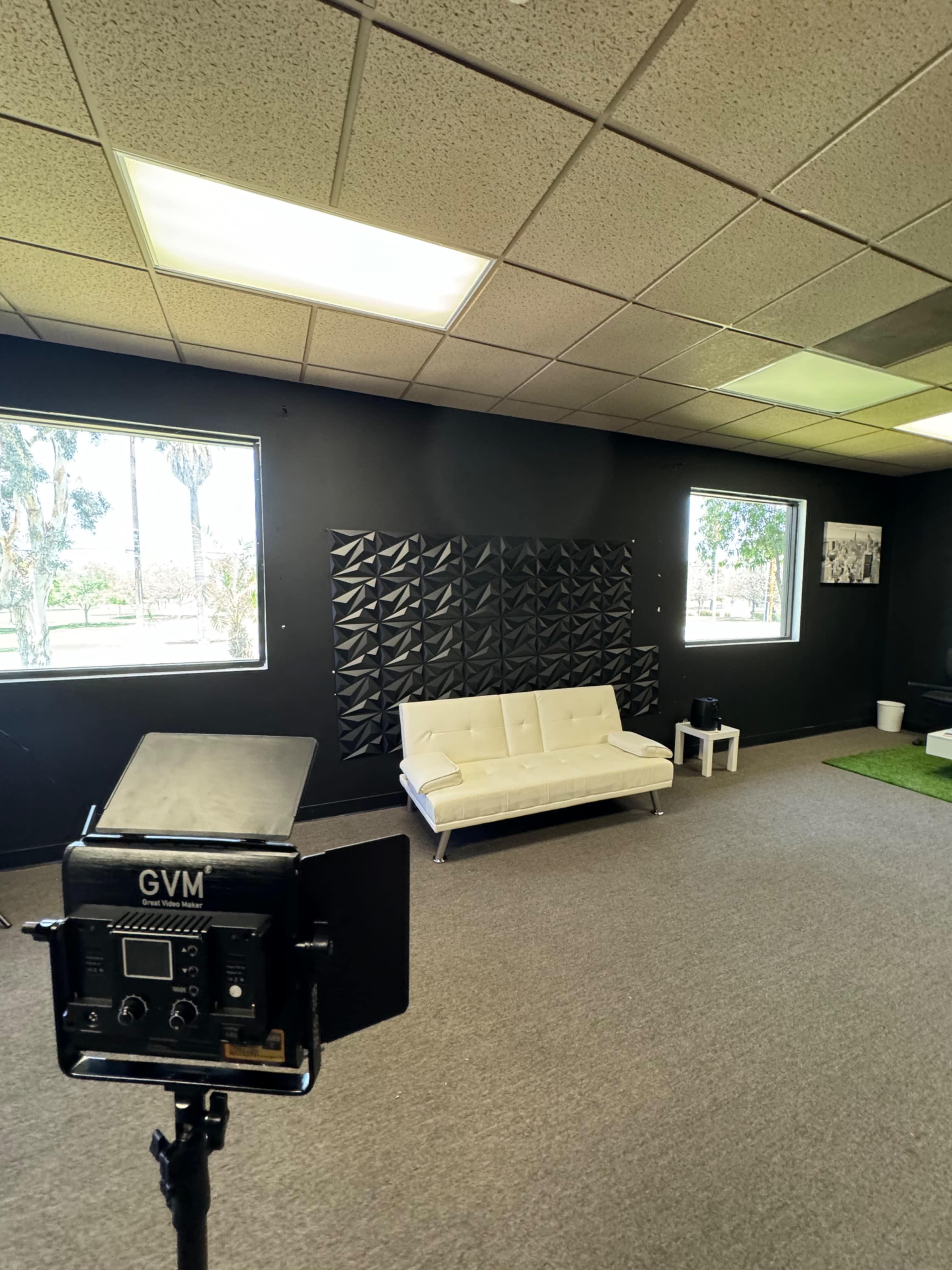 The image shows a minimalist studio space with a white couch, gray carpeting, and a black wall featuring soundproofing panels, all illuminated by natural light from windows.