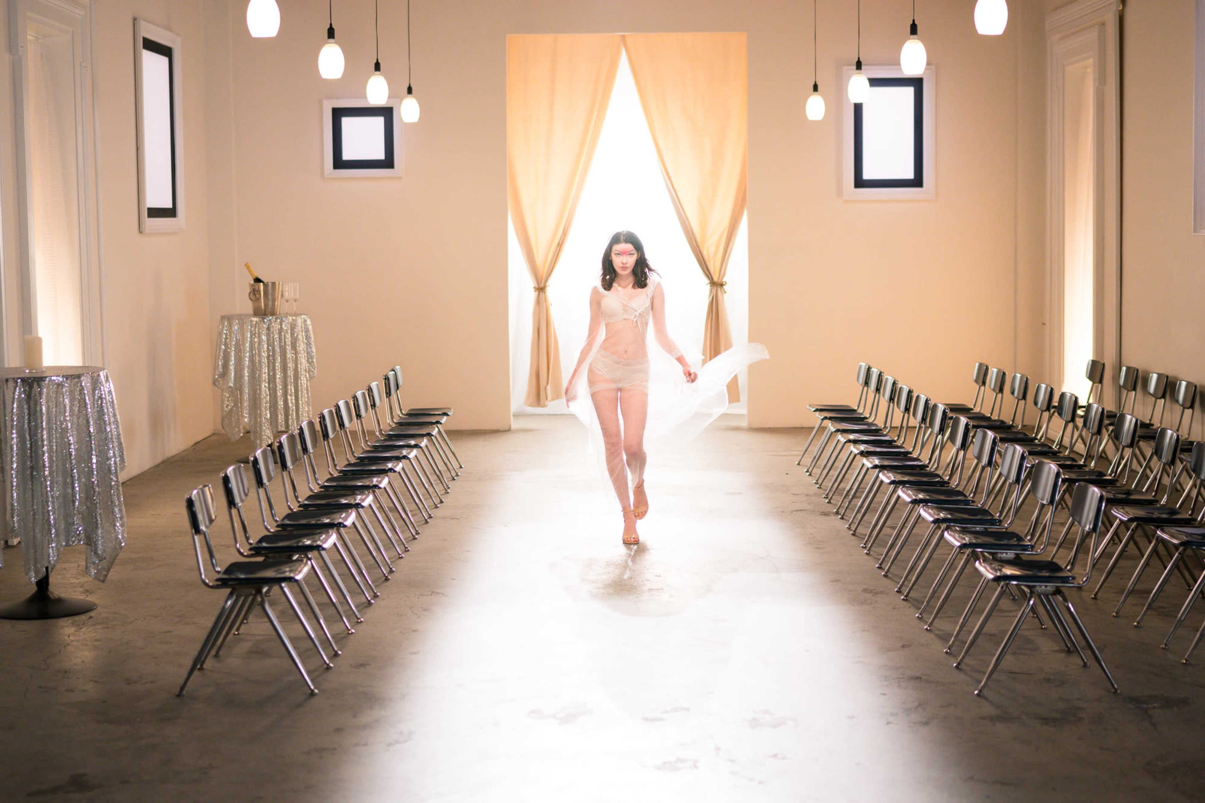 A model walks down a runway in a minimalist, well-lit space with rows of chairs on either side.