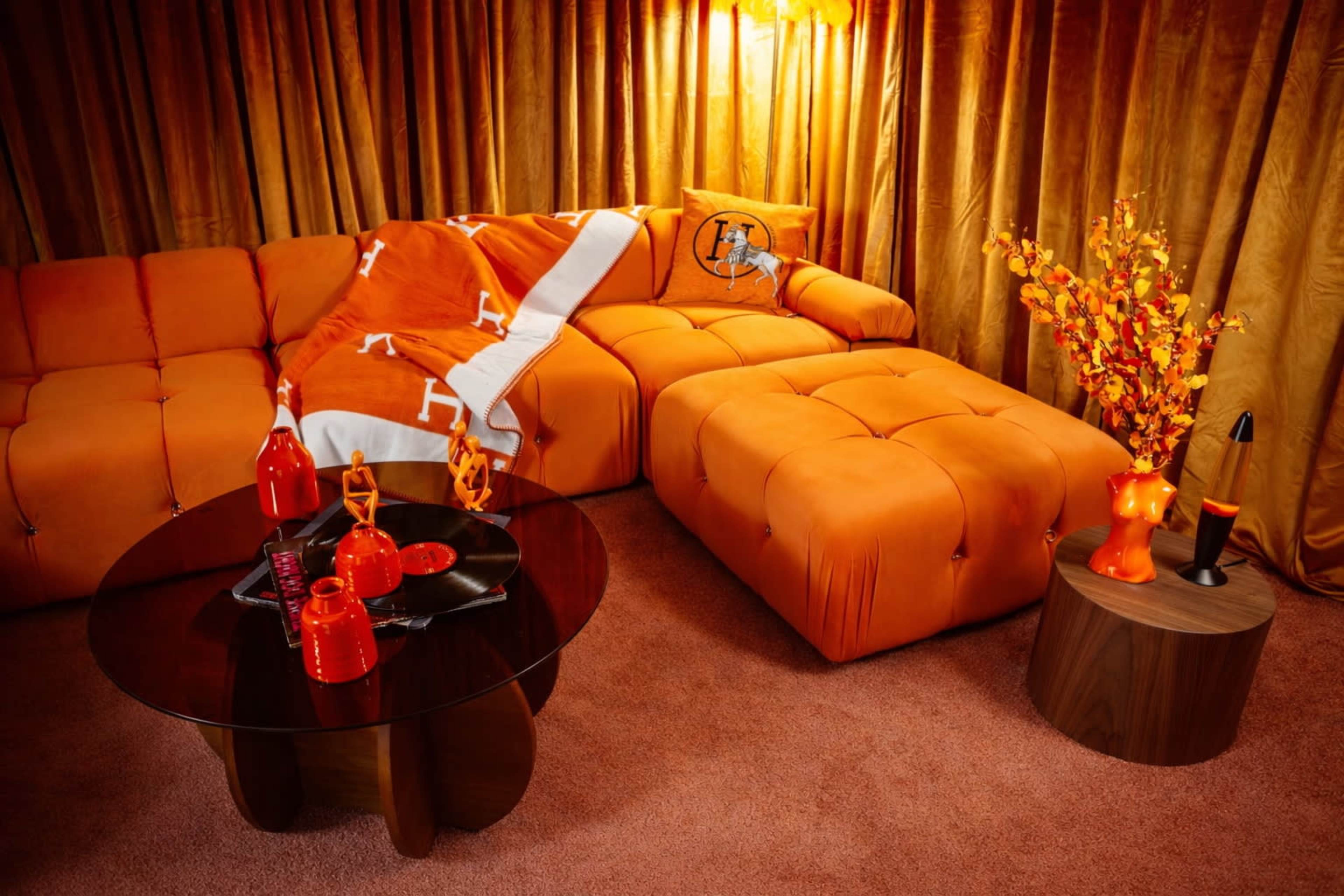 The image shows a cozy living room featuring an orange sectional sofa, a round glass coffee table, and decorative items in matching colors, set against golden curtains.