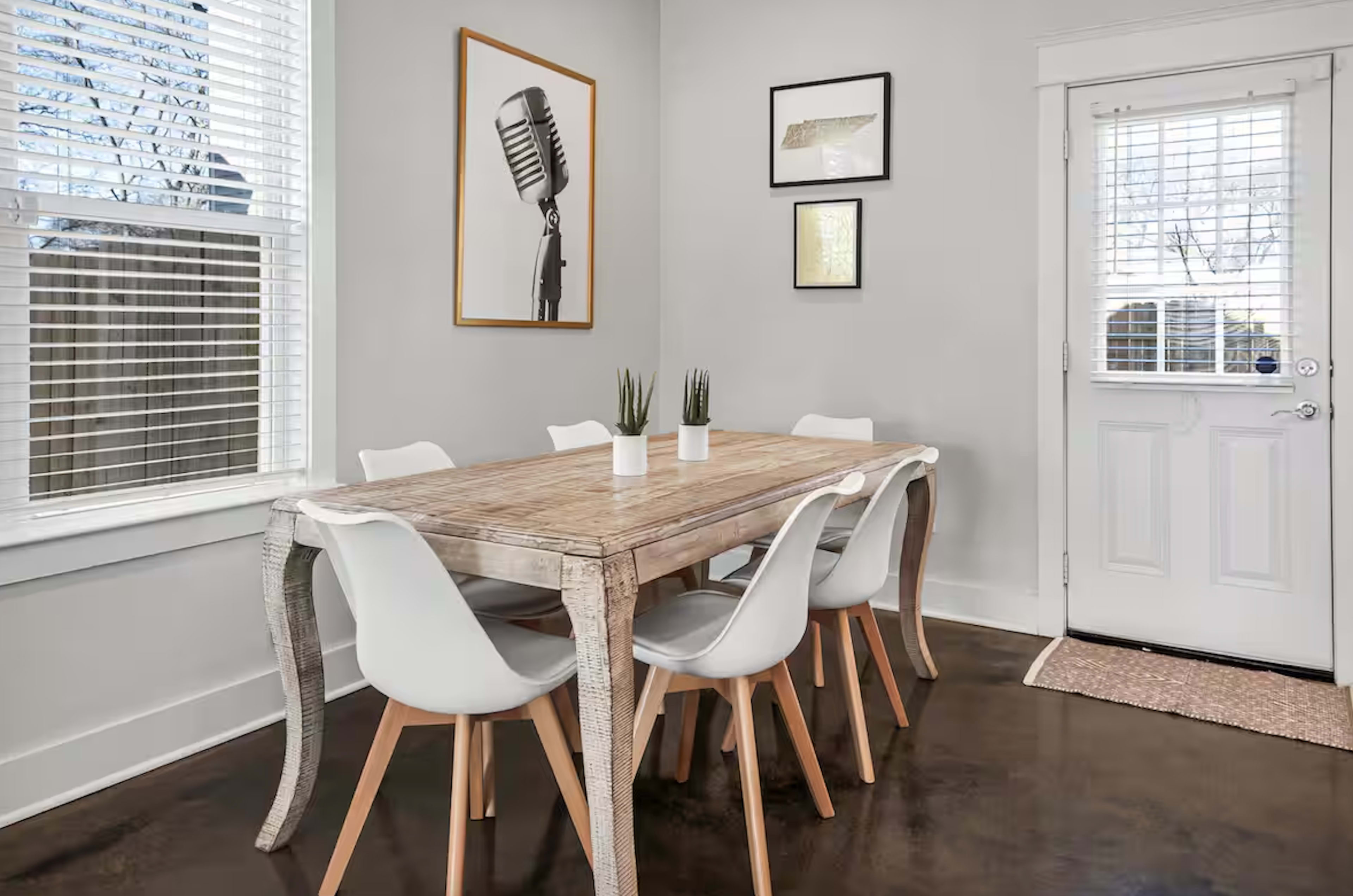 A dining area features a wooden table surrounded by six white chairs, with a microphone framed on the wall and a door leading outside.