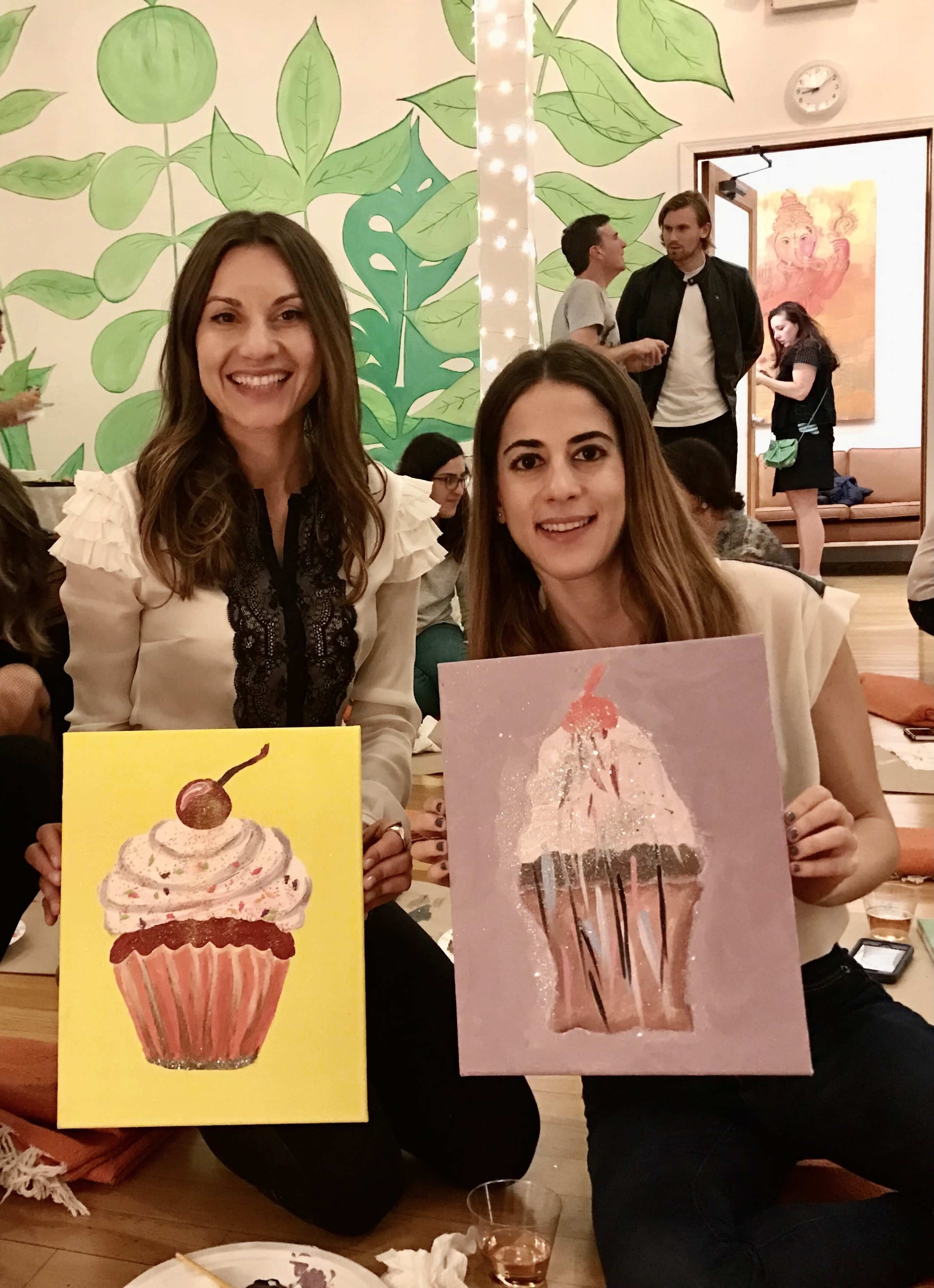 Two women hold up their painted canvas artworks of cupcakes in a vibrant, indoor setting with a green leafy mural on the wall.