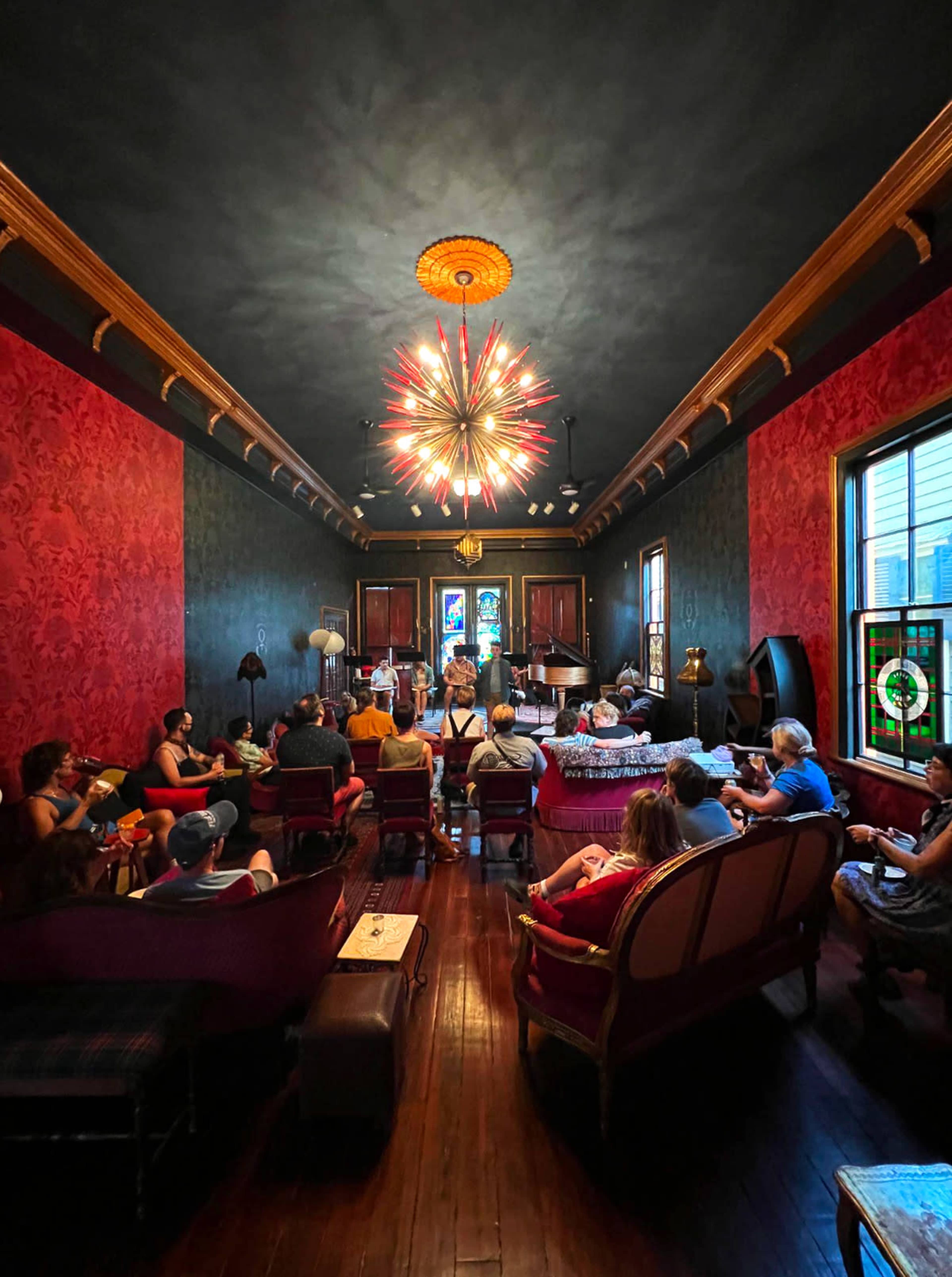 A group of people sit in a vintage-style room with patterned wallpaper and a large, modern chandelier, focused on a performer in front of them.
