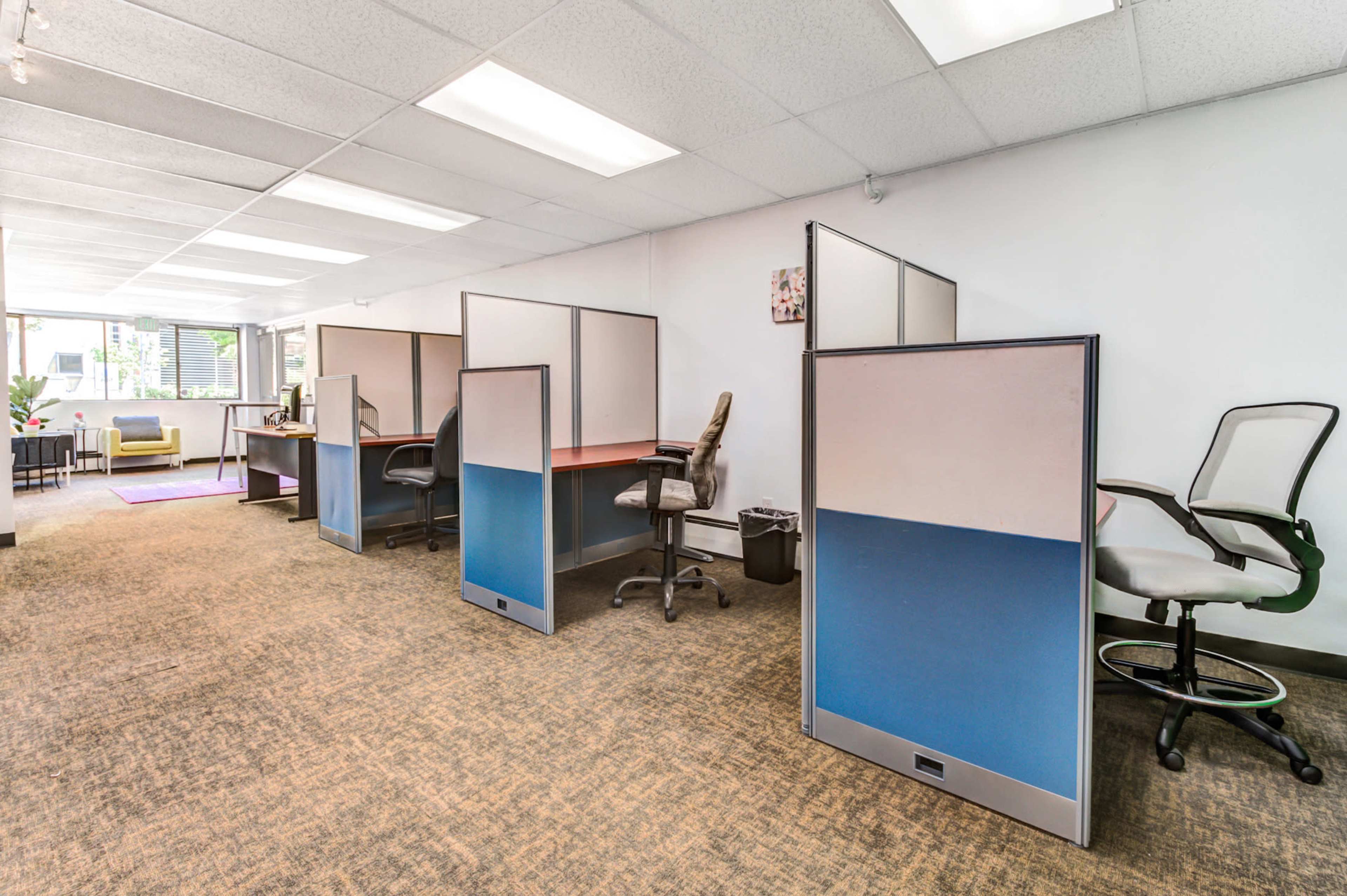 A spacious office layout with cubicles and ergonomic chairs arranged along one side, a seating area at the far end, and large windows allowing natural light to enter.