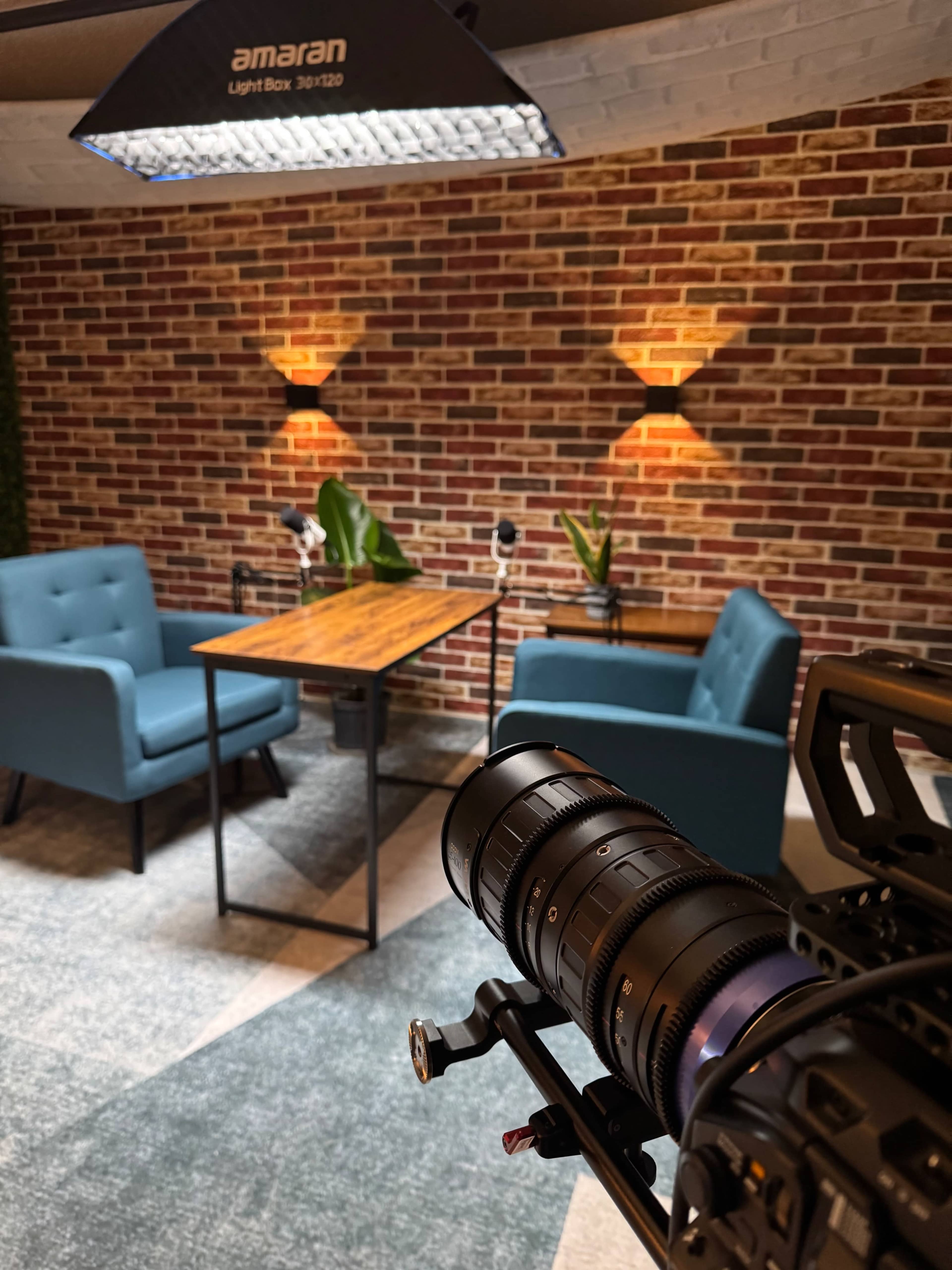 A camera is focused on a table with two blue chairs in a well-lit room featuring a brick wall and plants.