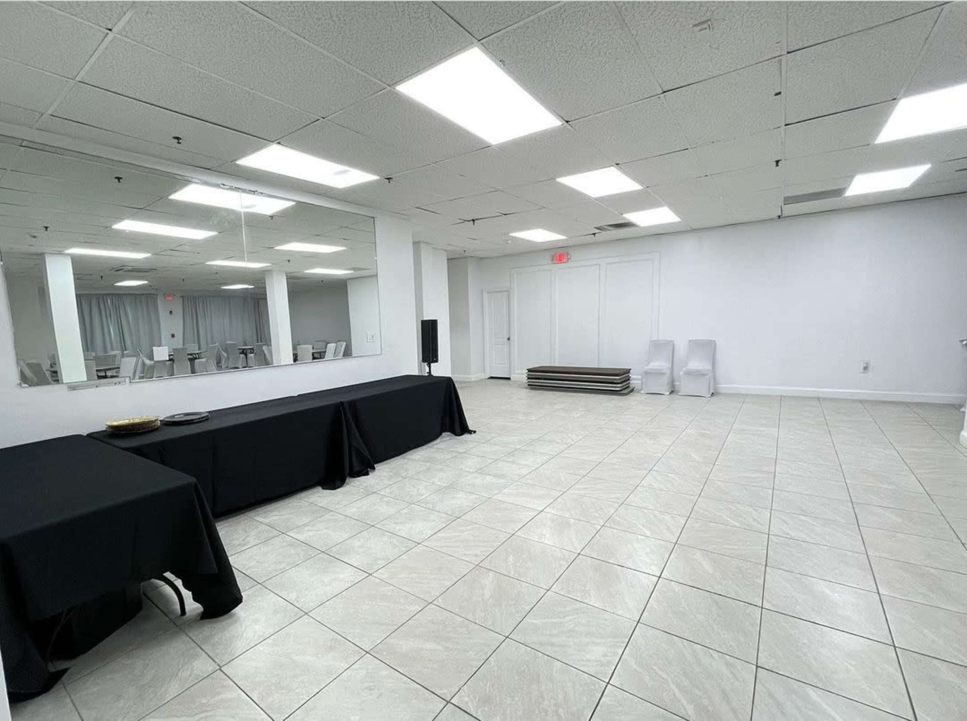 The image shows a spacious, empty room with tiled flooring, featuring a long black table, a large wall mirror, and a few white chairs arranged against the wall.