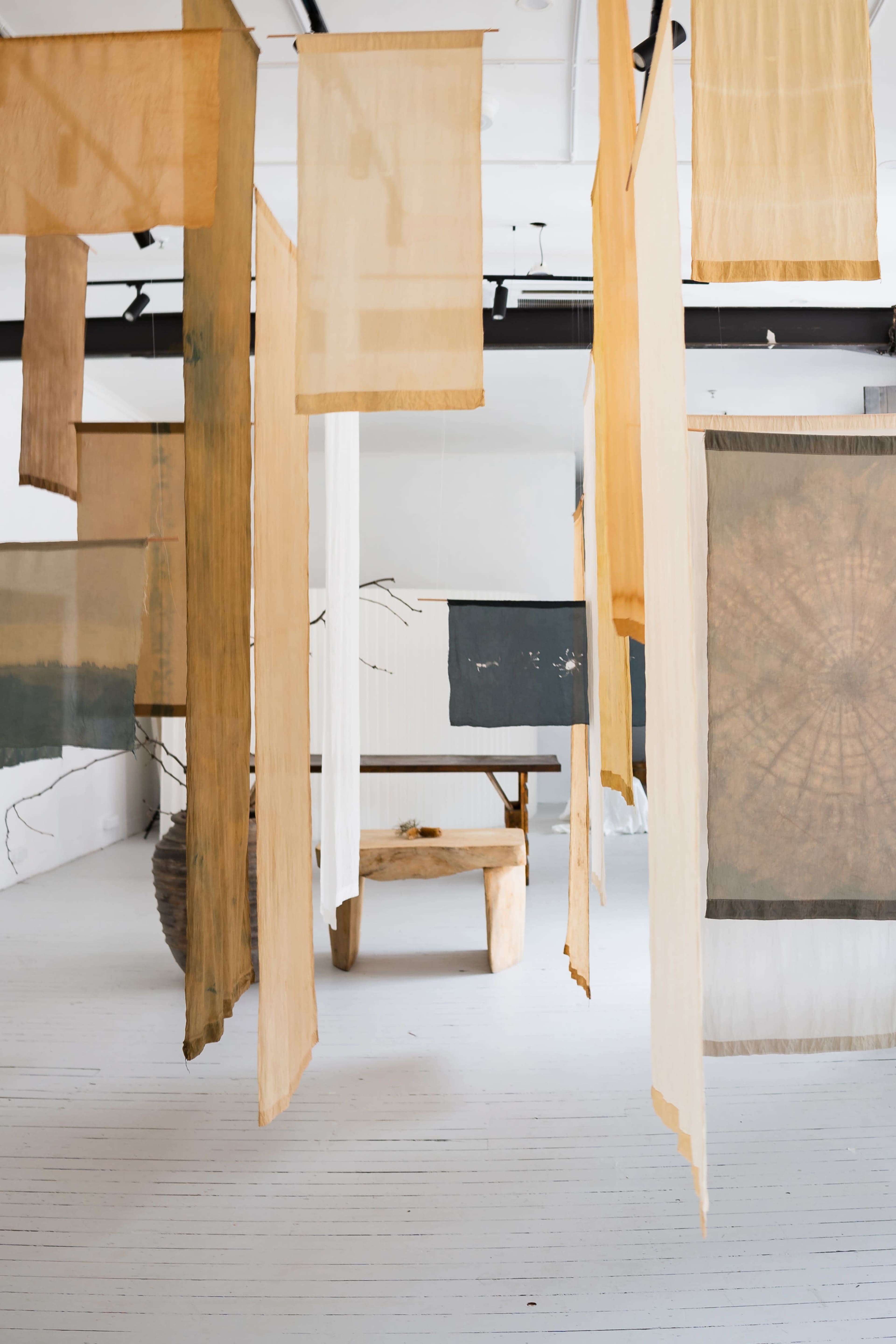 The image shows a spacious art gallery with suspended fabric panels of various colors and patterns, arranged between wooden furniture and an empty floor.