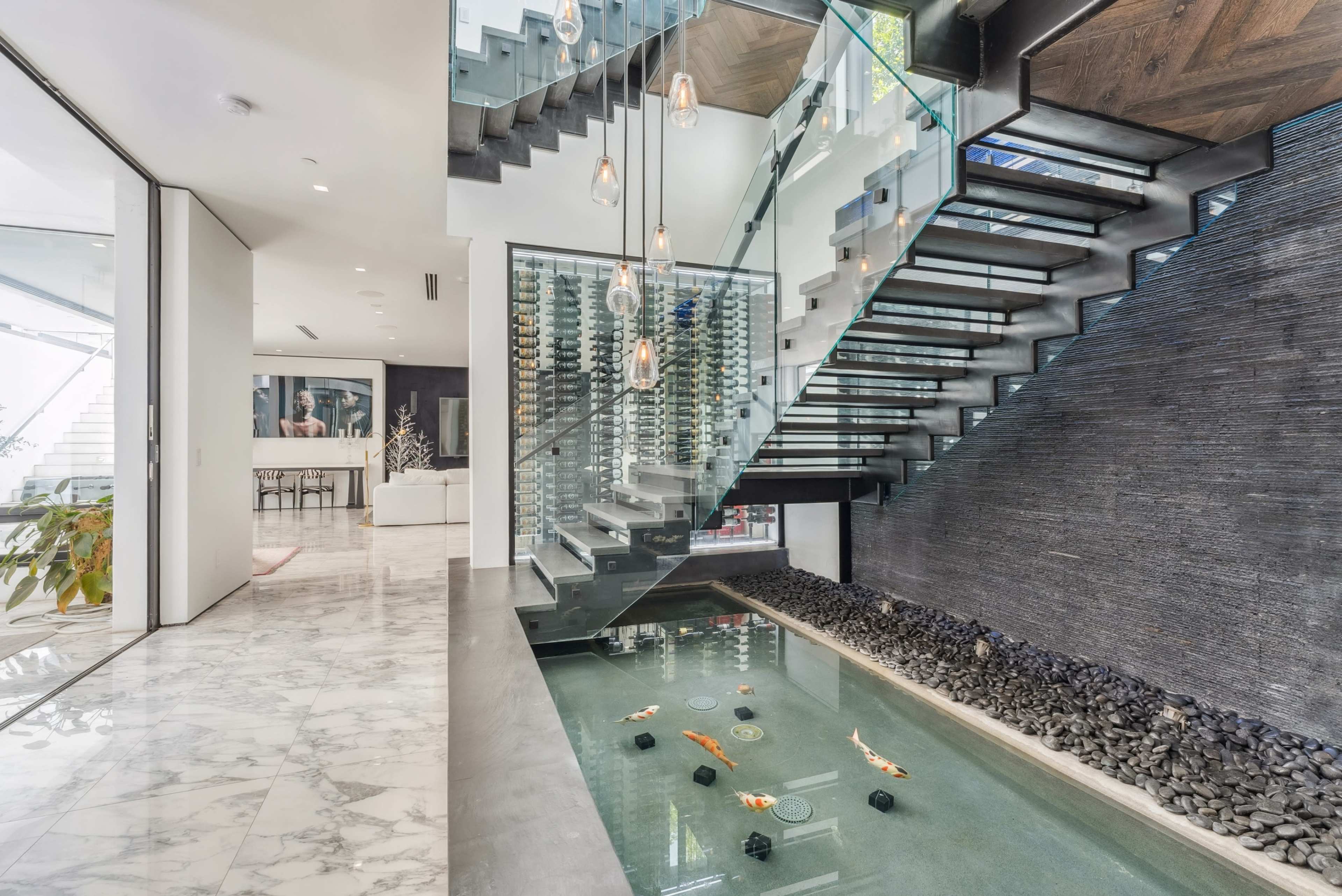 The image features a modern entryway with a glass staircase, a water feature containing koi fish, and sleek, minimalist design elements.